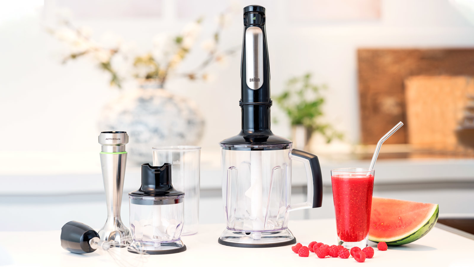 A black and silver immersion blender and accessories are on a white surface with a glass of red drink.