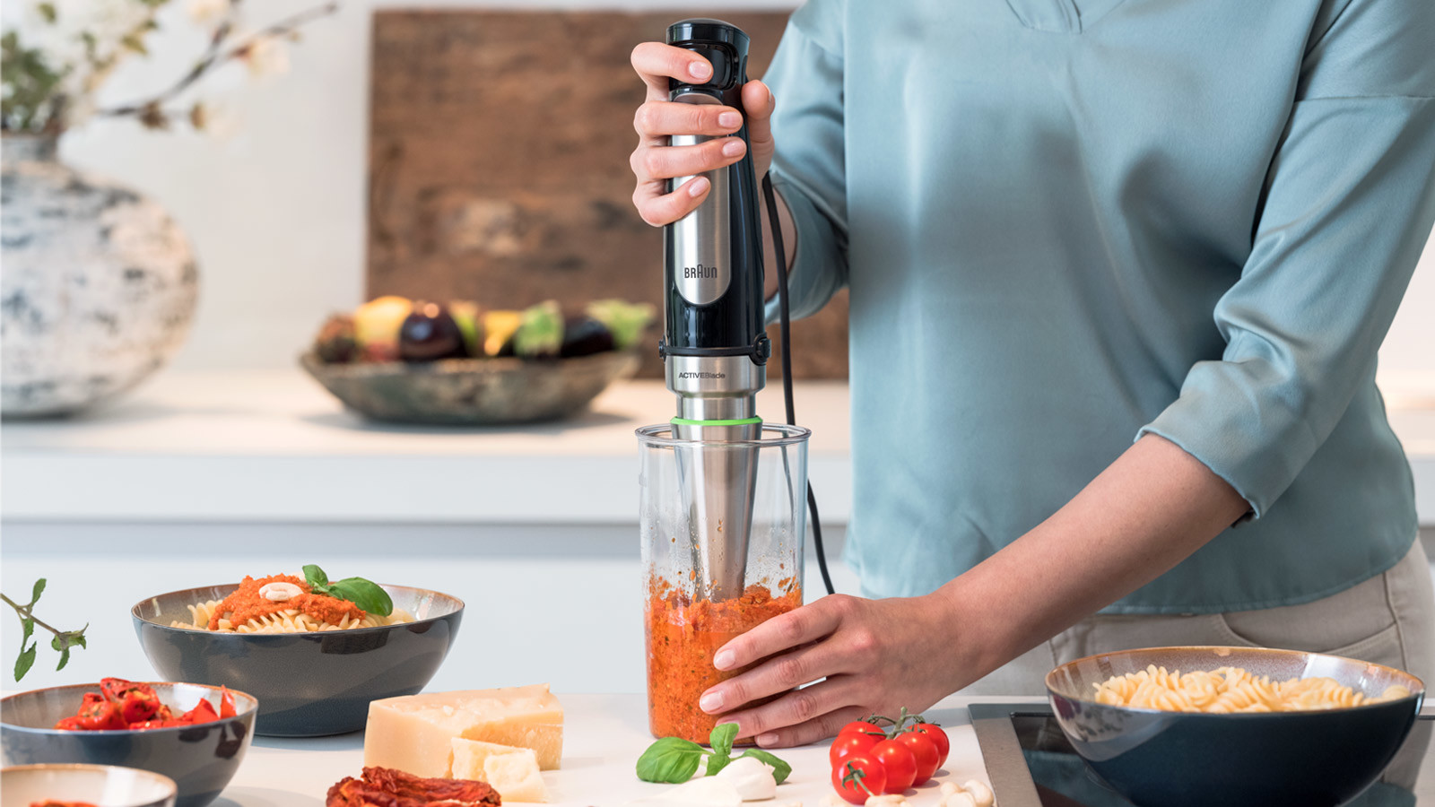 Woman blending ingredients with a hand blender in a kitchen. Various ingredients around.