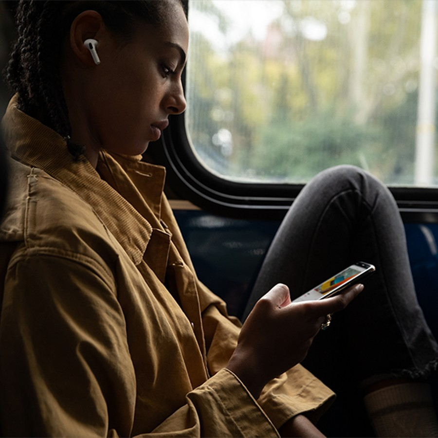 Mujer en chaqueta usando un teléfono, con auriculares, sentada en un tren. La ventana está detrás.