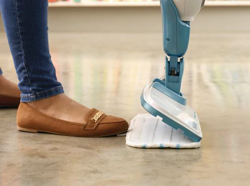 Person's feet in brown shoes and jeans with a steam mop in action on a floor.