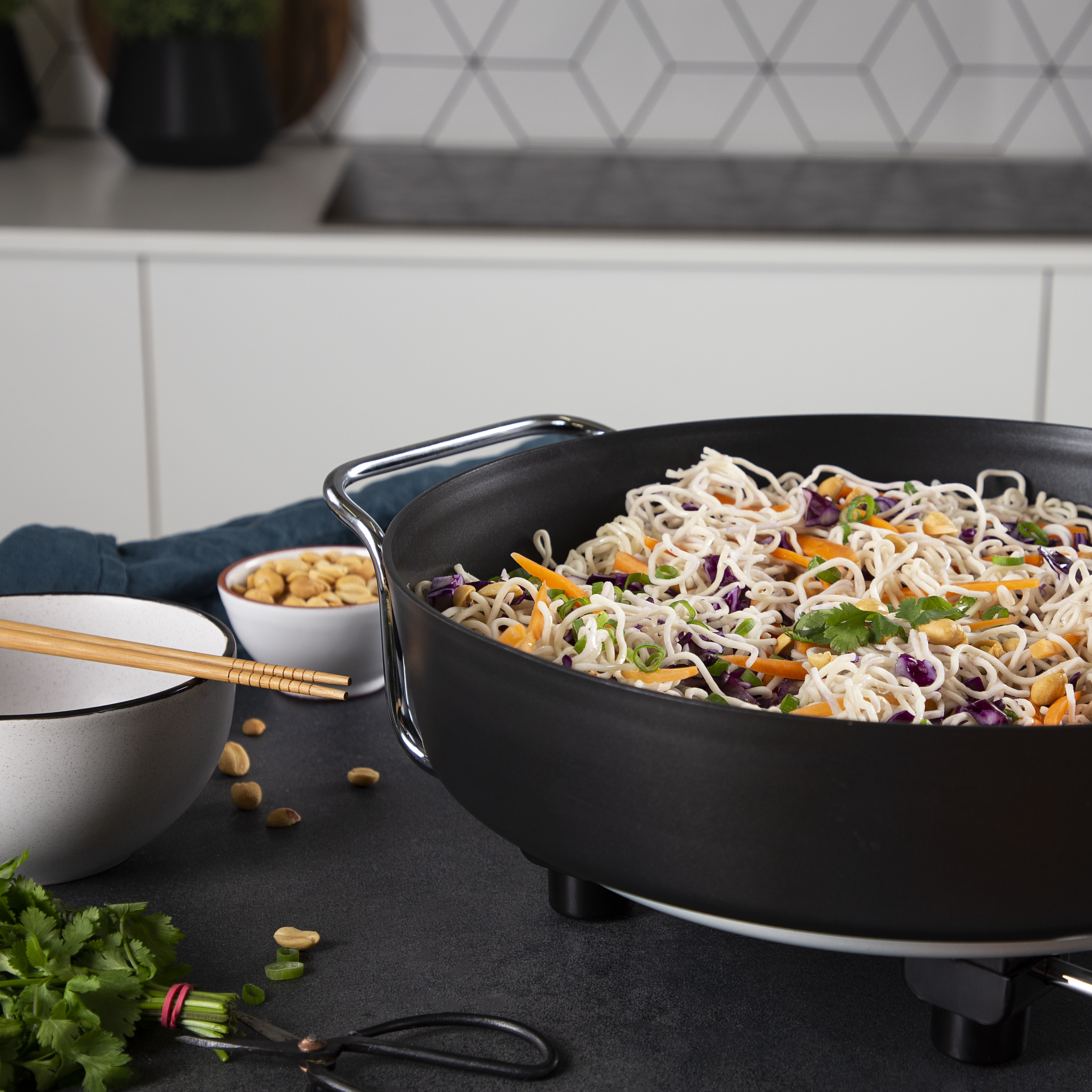 Sartén con fideos, verduras y cacahuetes. Palillos chinos y un cuenco también presentes.