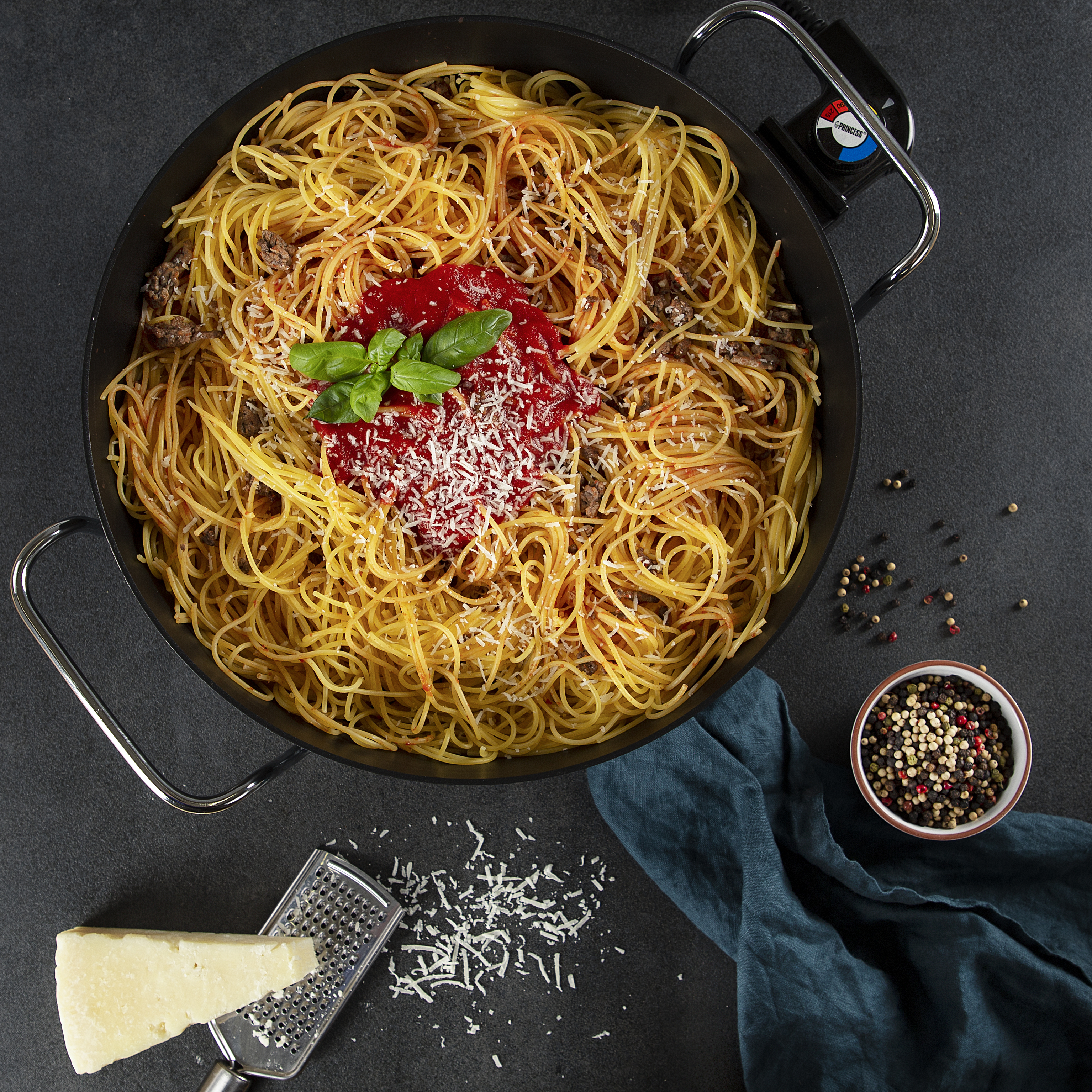 Espagueti con salsa de tomate, queso y albahaca en una sartén, con ingredientes alrededor.