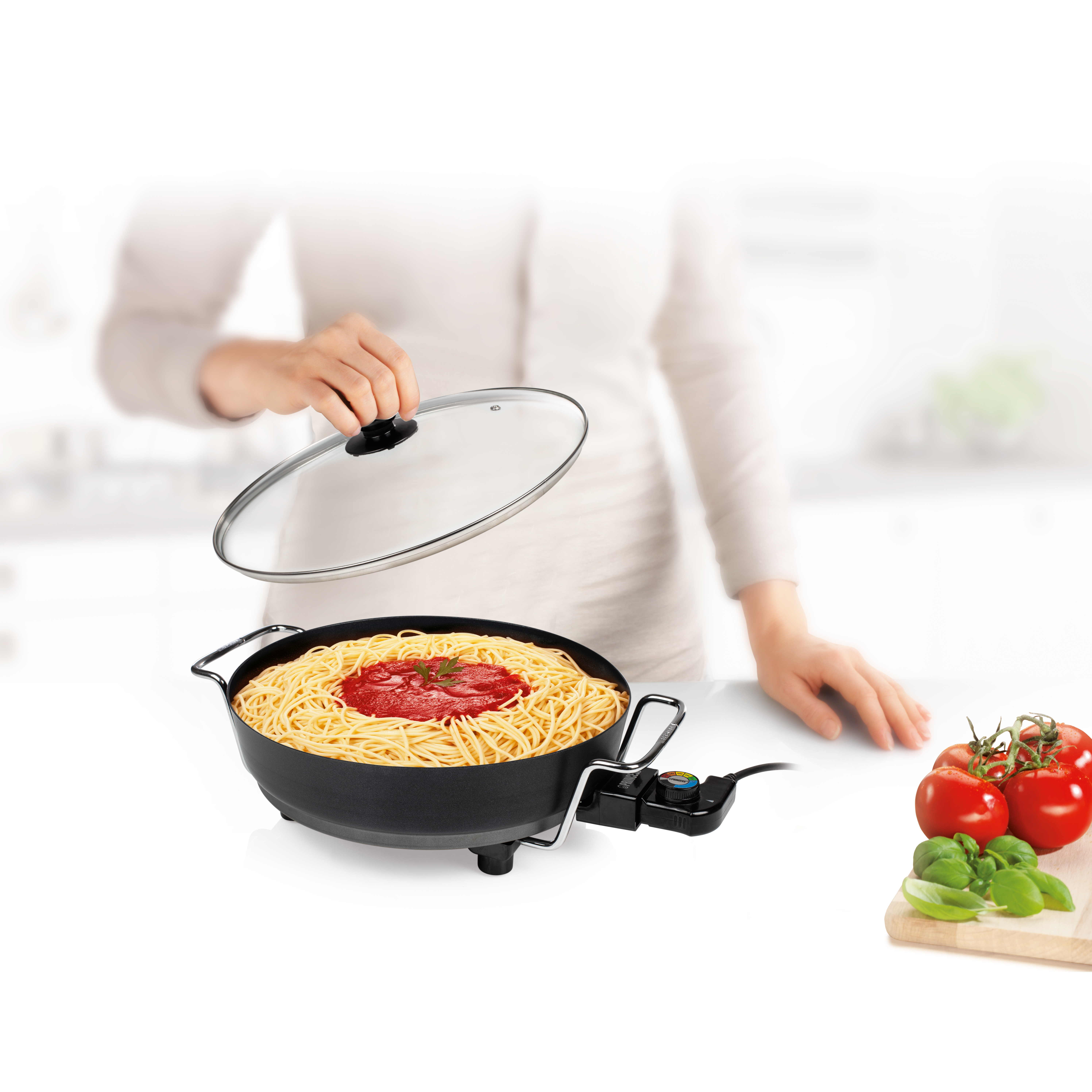 Una persona sosteniendo una tapa sobre una olla de espaguetis con salsa. Tomates y albahaca a un lado.