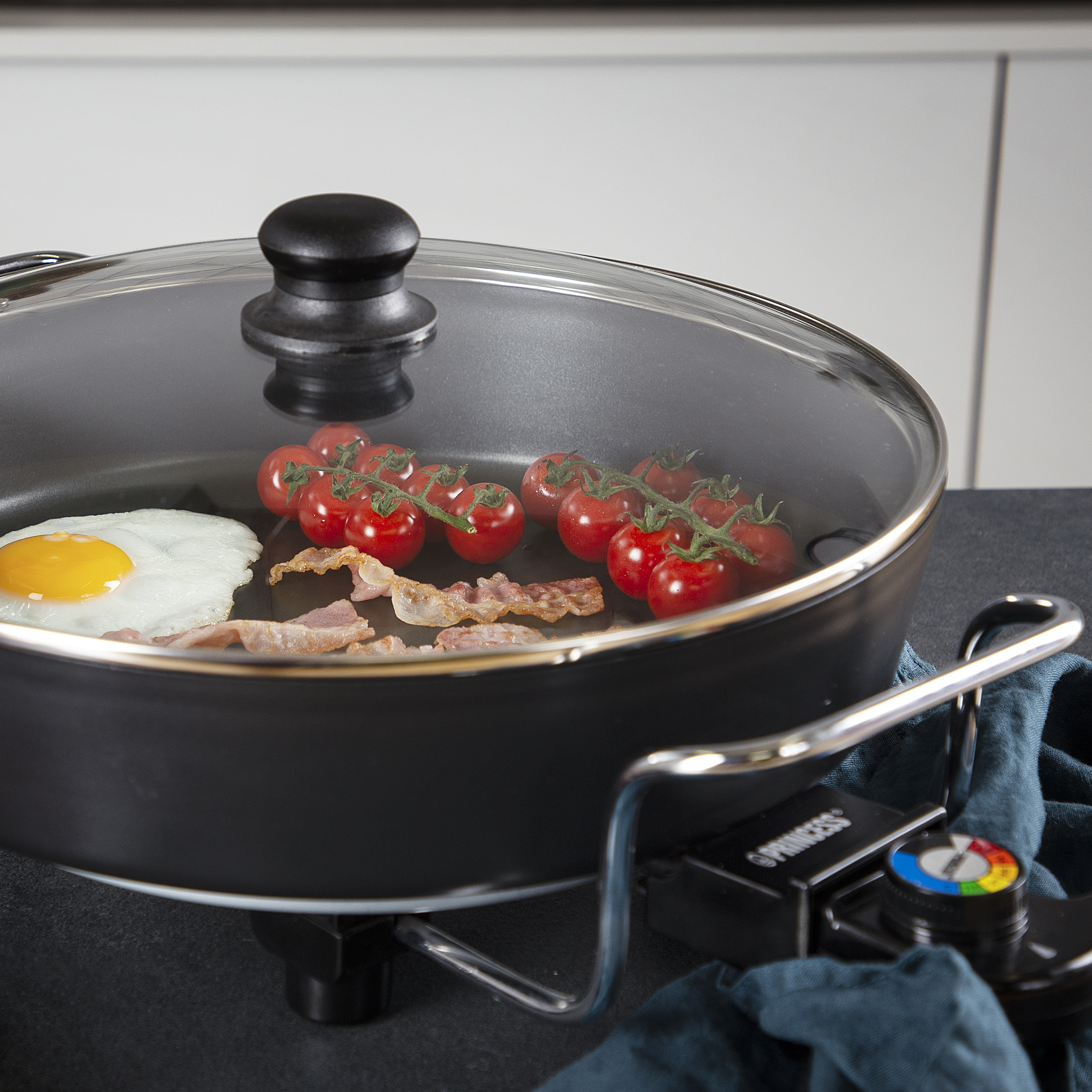 Sartén eléctrica con huevo, tocino y tomates, con tapa de vidrio.