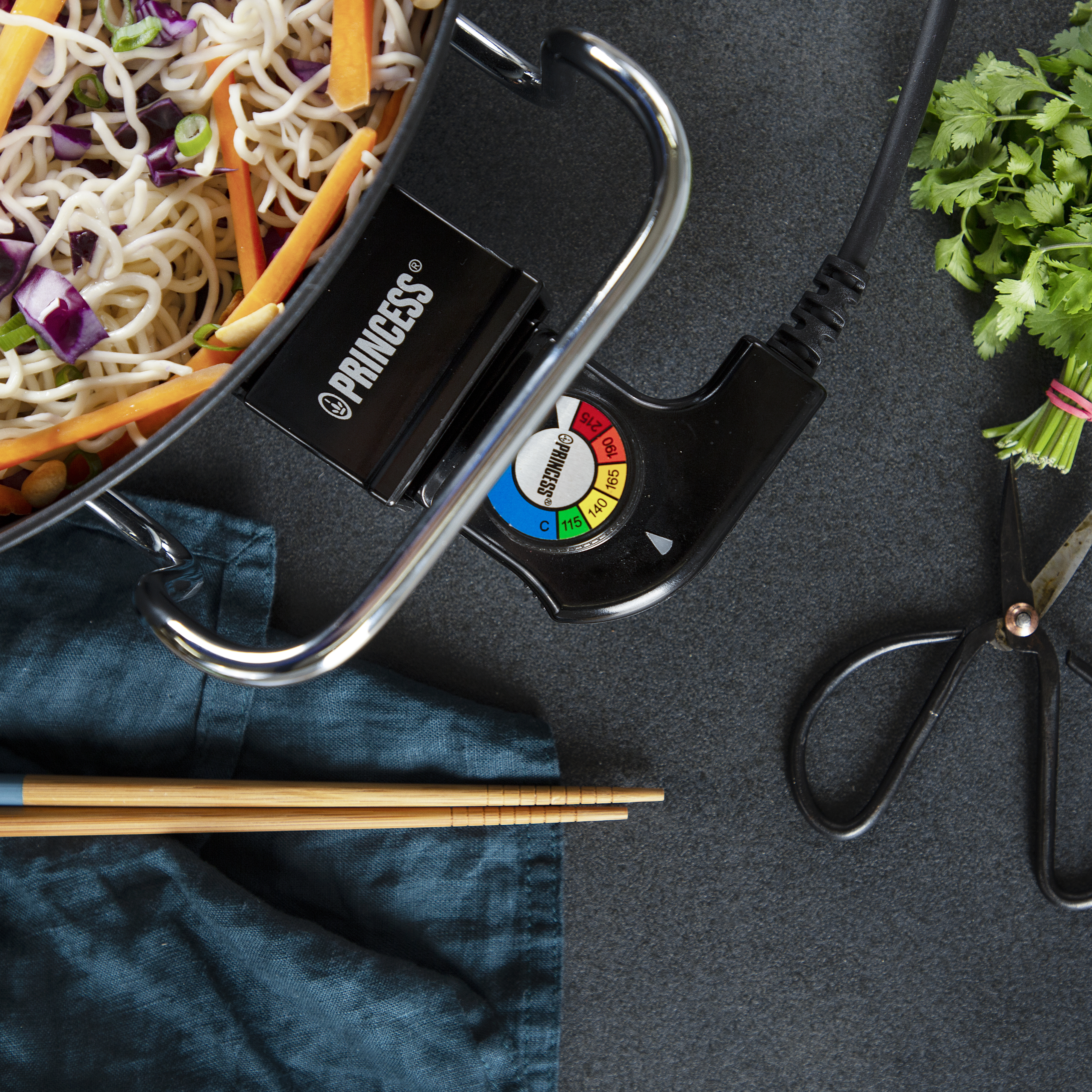 Ingredientes para comida asiática: fideos, verduras y cilantro junto a sartén y tijeras.