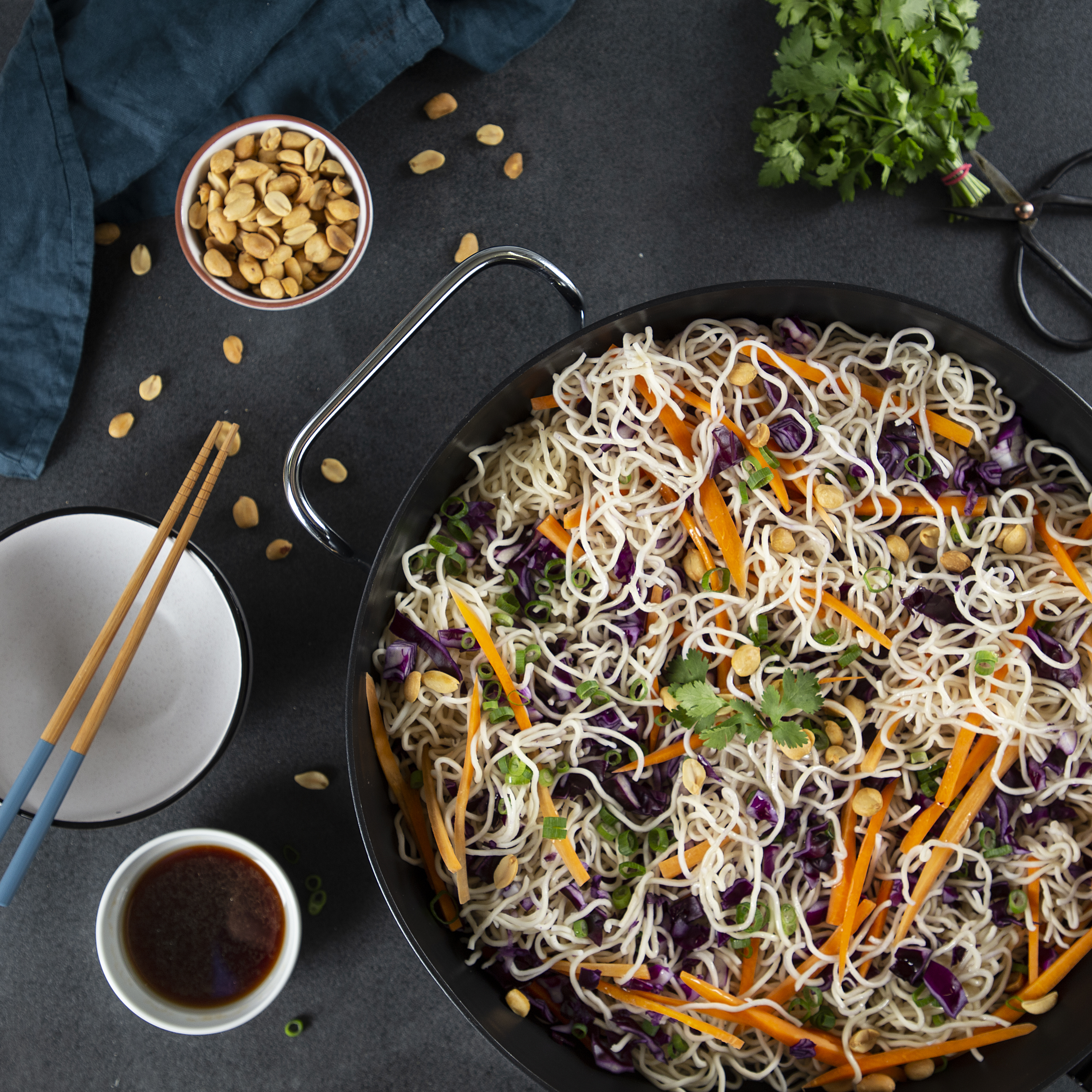 Una sartén con fideos, verduras y cacahuetes, servida con palillos y salsa.