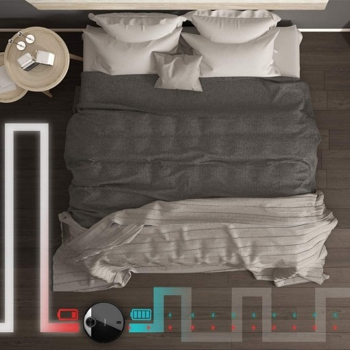 Una vista superior de una cama con una aspiradora robot. La aspiradora es negra. El suelo es de madera.