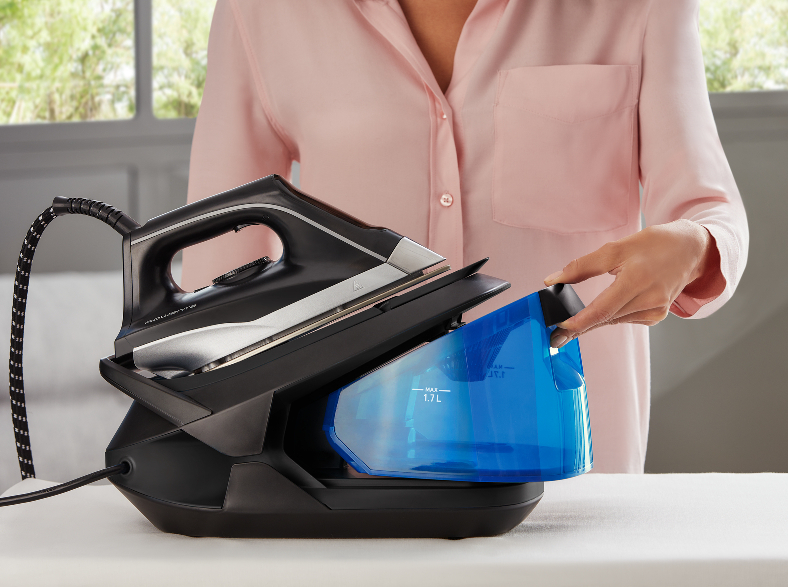Mujer llenando tanque de agua azul de plancha, superficie blanca y ventana.