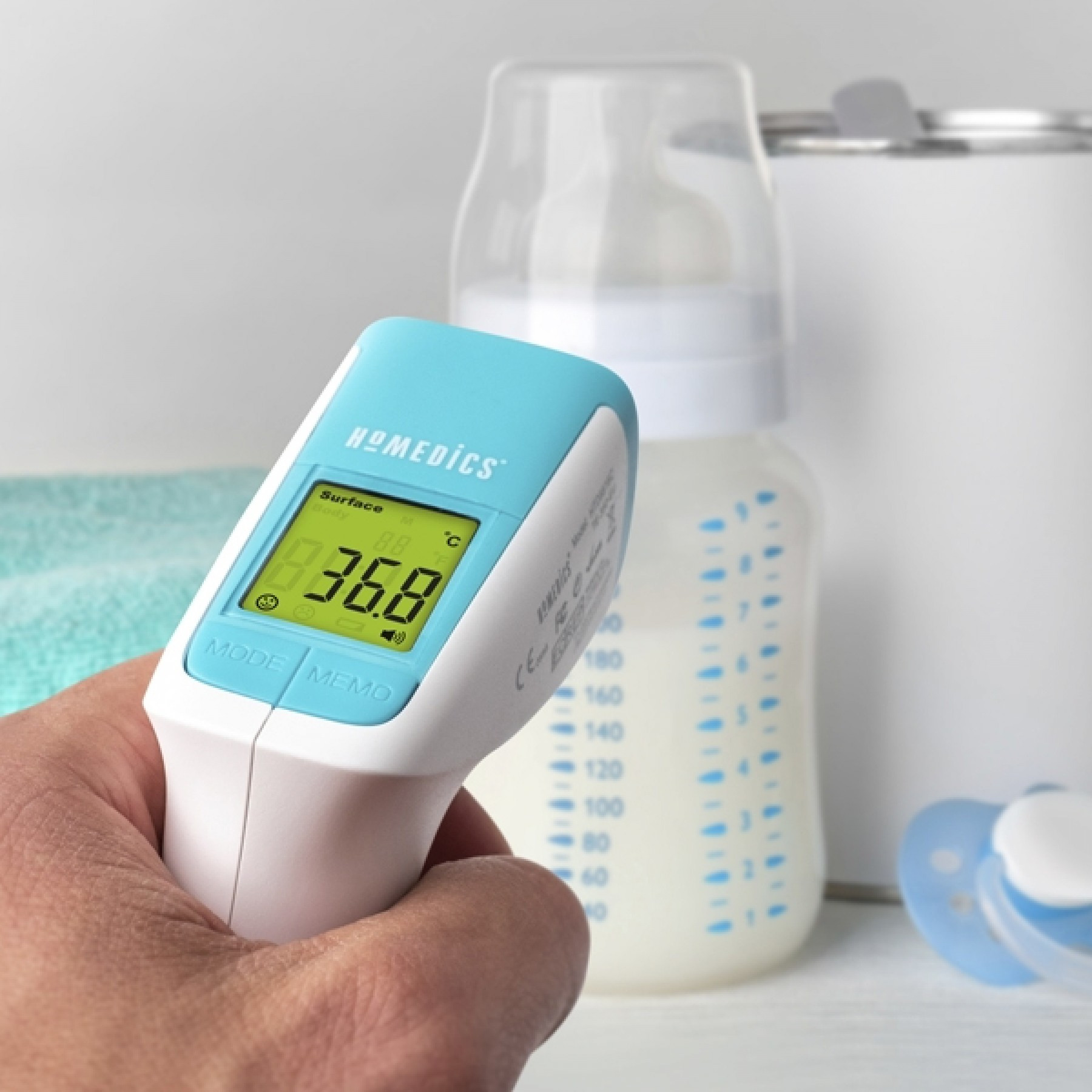 Een hand die een thermometer vasthoudt, leest 36,8°C af. Een fles en een fopspeen op de achtergrond.