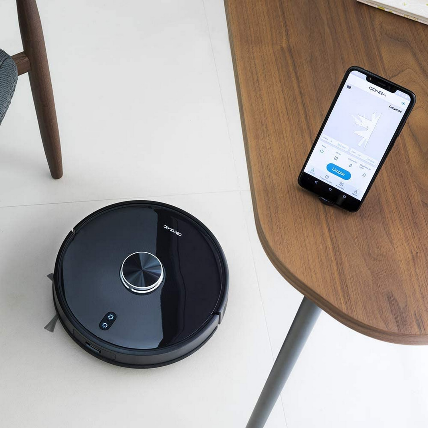 Una aspiradora robótica negra y un teléfono en una mesa de madera, mostrando la interfaz de la app.