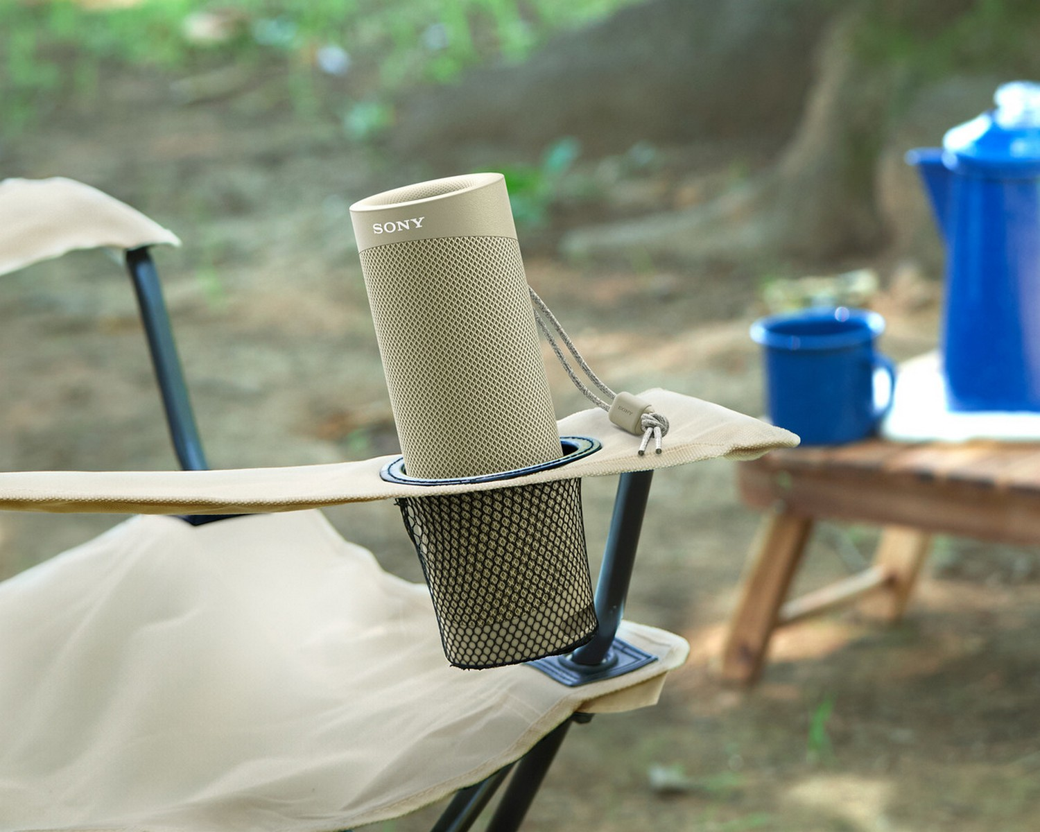 Un altavoz beige en un portavasos de silla con una taza azul y una tetera azul en el fondo, al aire libre.