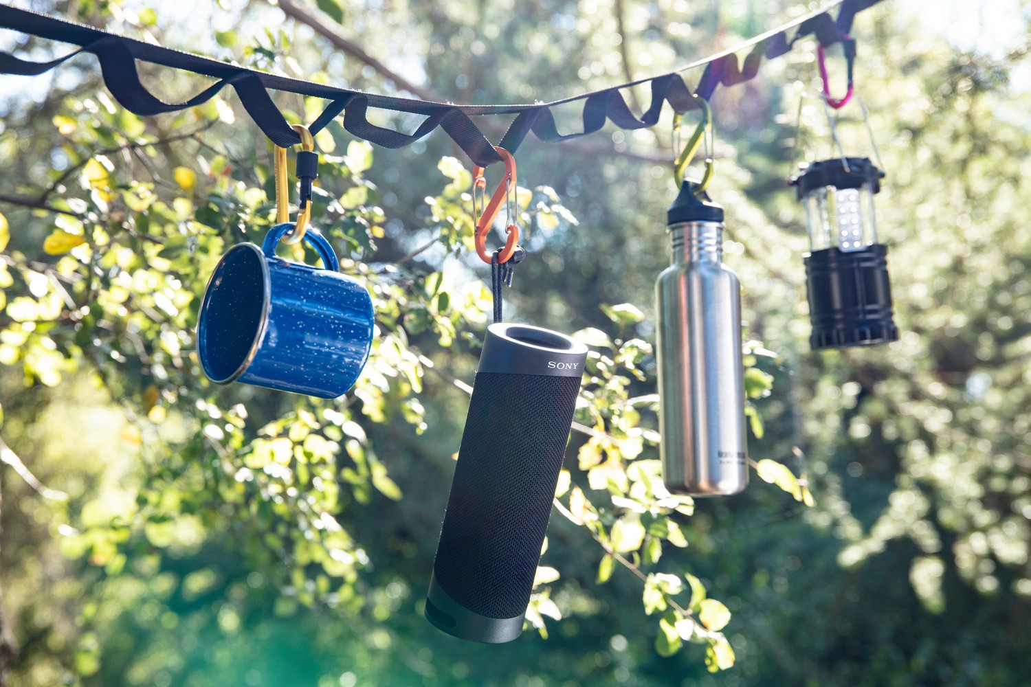 Escena de acampada con taza, altavoz, botella de agua y linterna colgando de una cuerda frente a árboles.