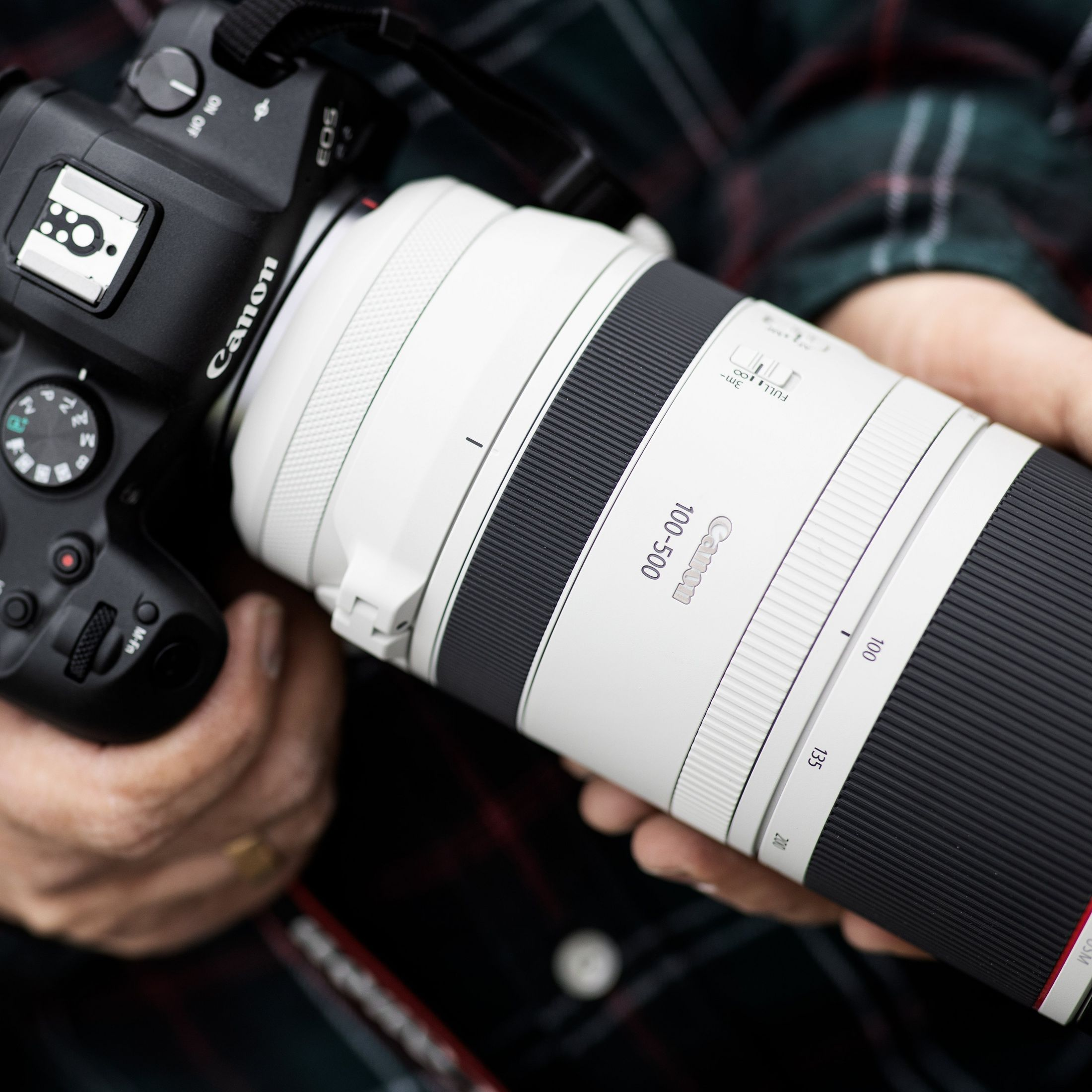 Una persona tiene in mano una fotocamera Canon con un lungo obiettivo bianco. L'obiettivo dice Canon 100-500.