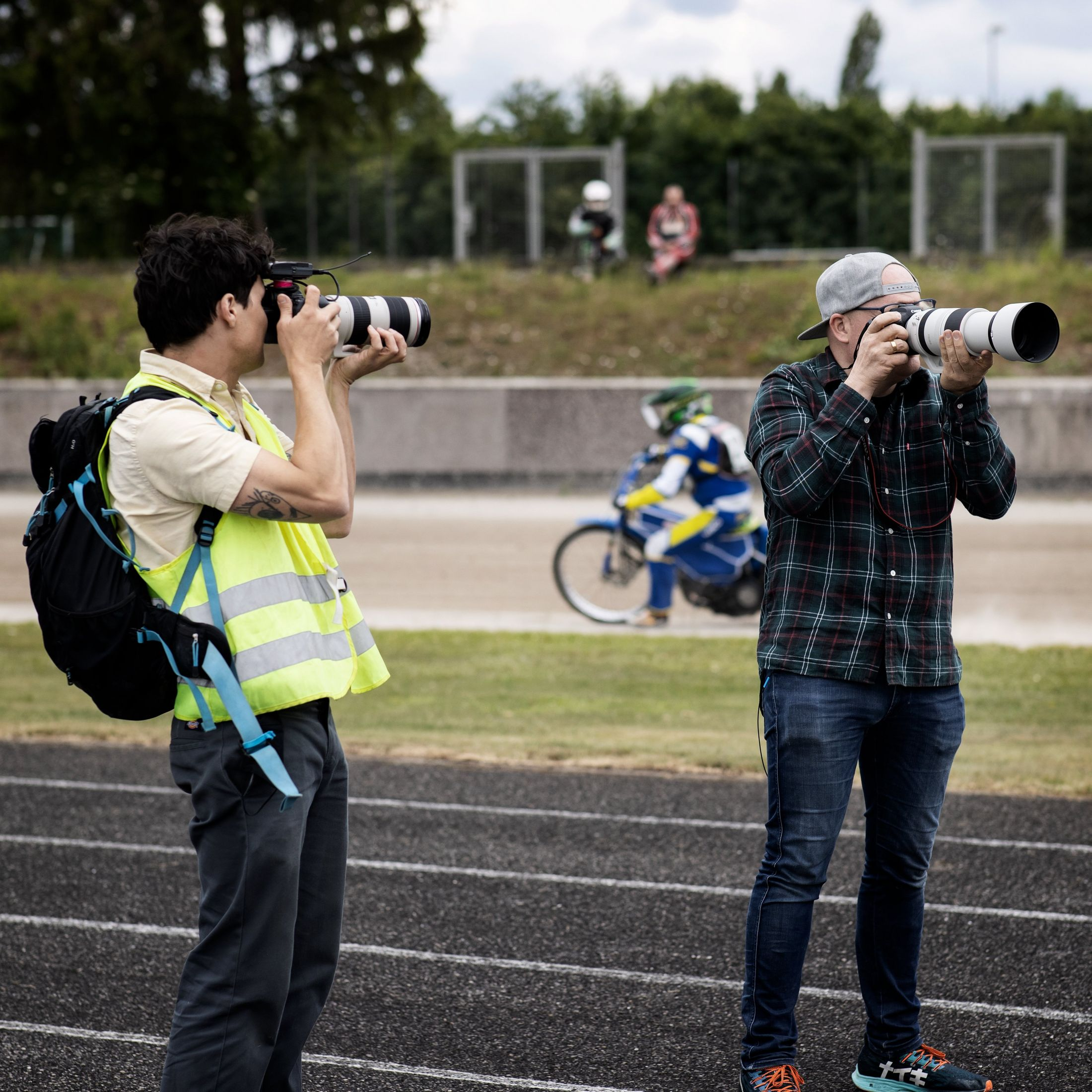 Due fotografi con macchine fotografiche, uno con un gilet luminoso. Piloti di moto su una pista sullo sfondo.