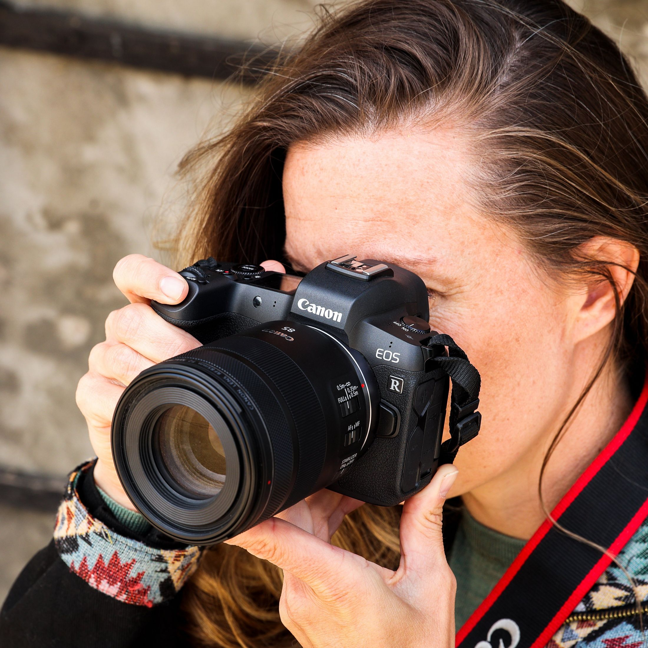Una donna che tiene una fotocamera Canon EOS R, guardando attraverso il mirino. È all'aperto.