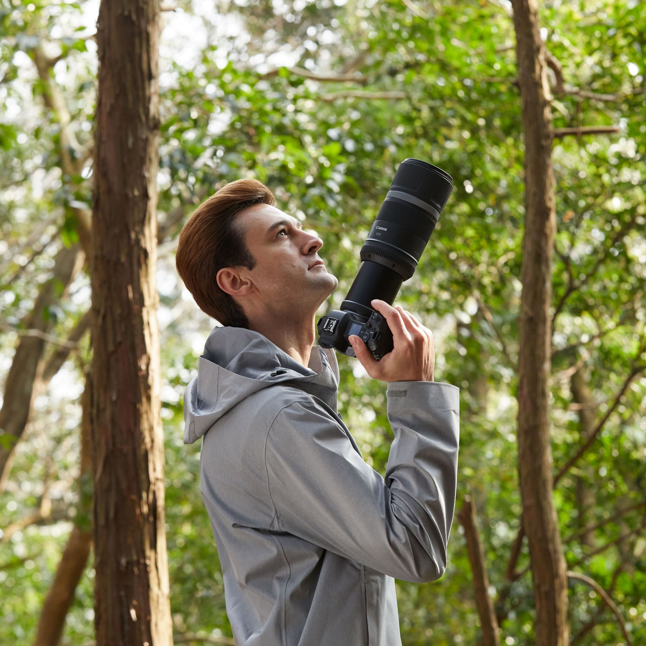 Un homme en veste grise tient un appareil photo avec objectif dans une forêt.