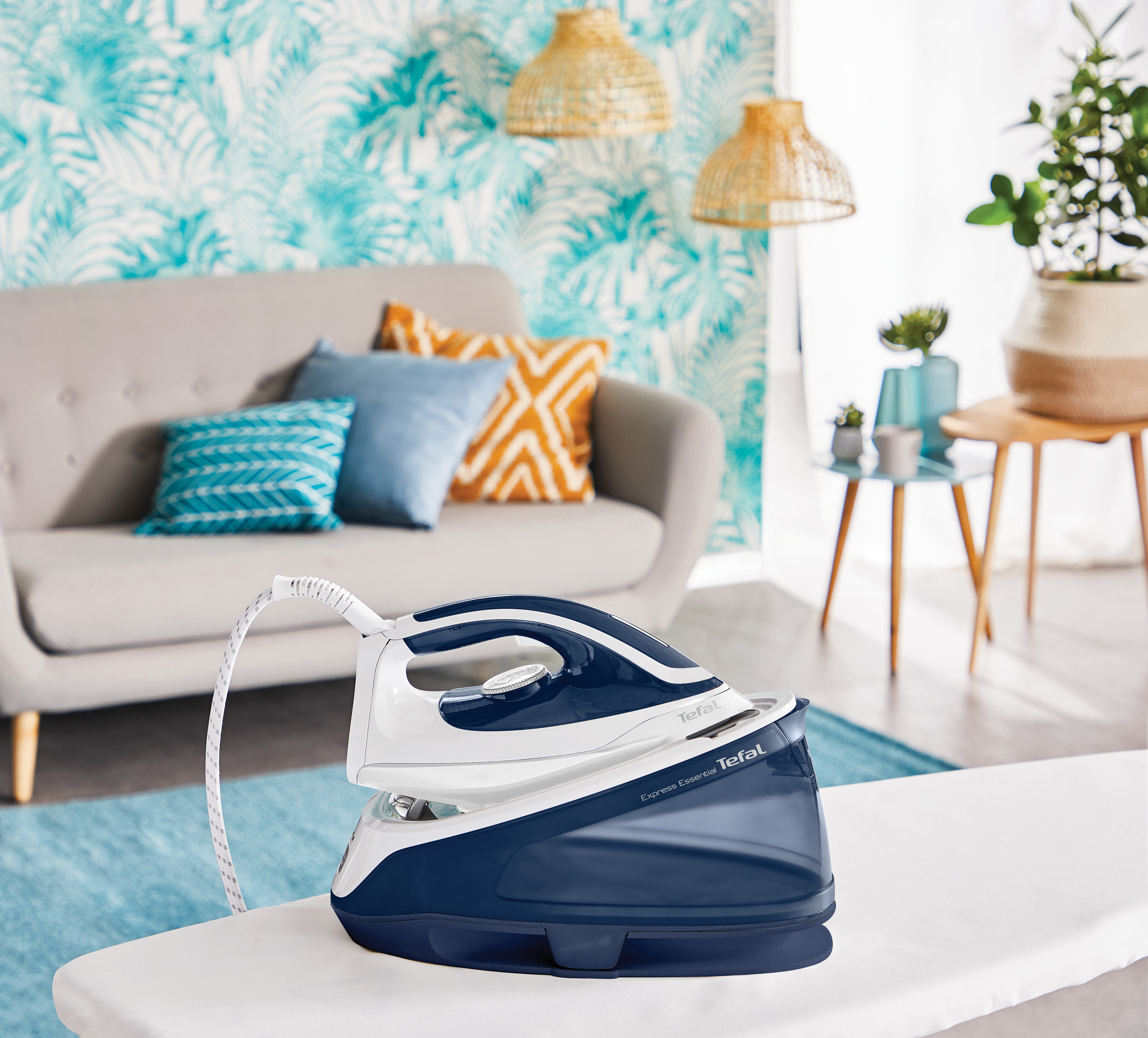 Bügeleisen auf Bügelbrett, blau und weiß. Graues Sofa, blaue Kissen und Deko im Hintergrund.