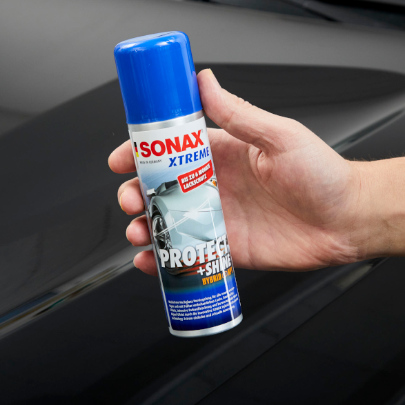 Kéz kék-fehér spray-t tart fekete autó felett, ragyogásért és védelemért.