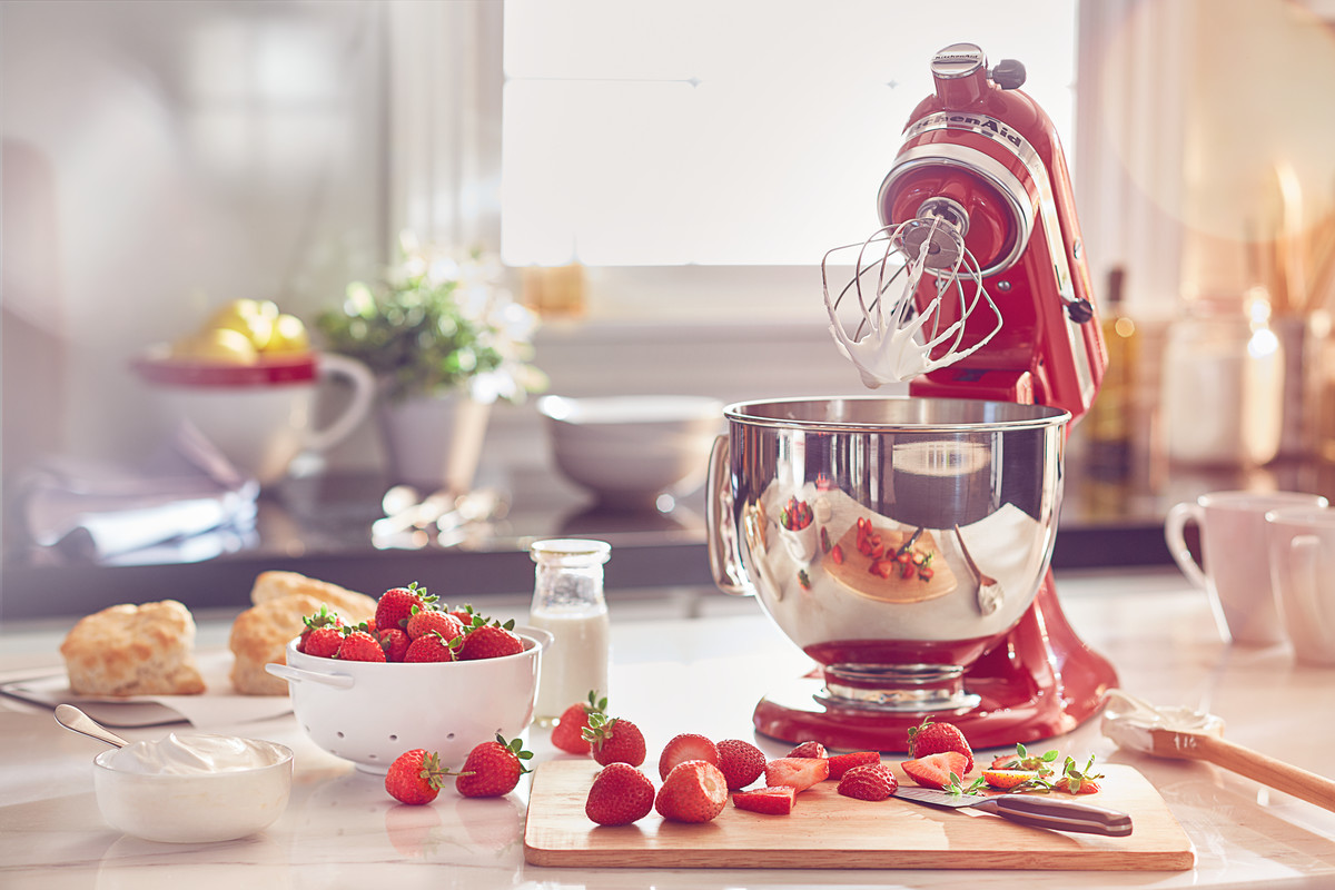 Ein roter KitchenAid-Mixer mit einer Schüssel mit Baiser, Erdbeeren und einem Schneidebrett auf einer Küchentheke.