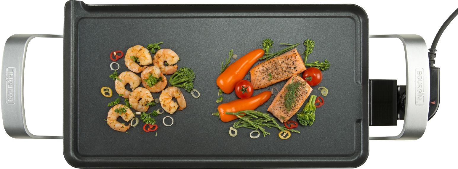 A griddle with grilled shrimp, salmon, and vegetables, with silver handles. It is on a black surface.