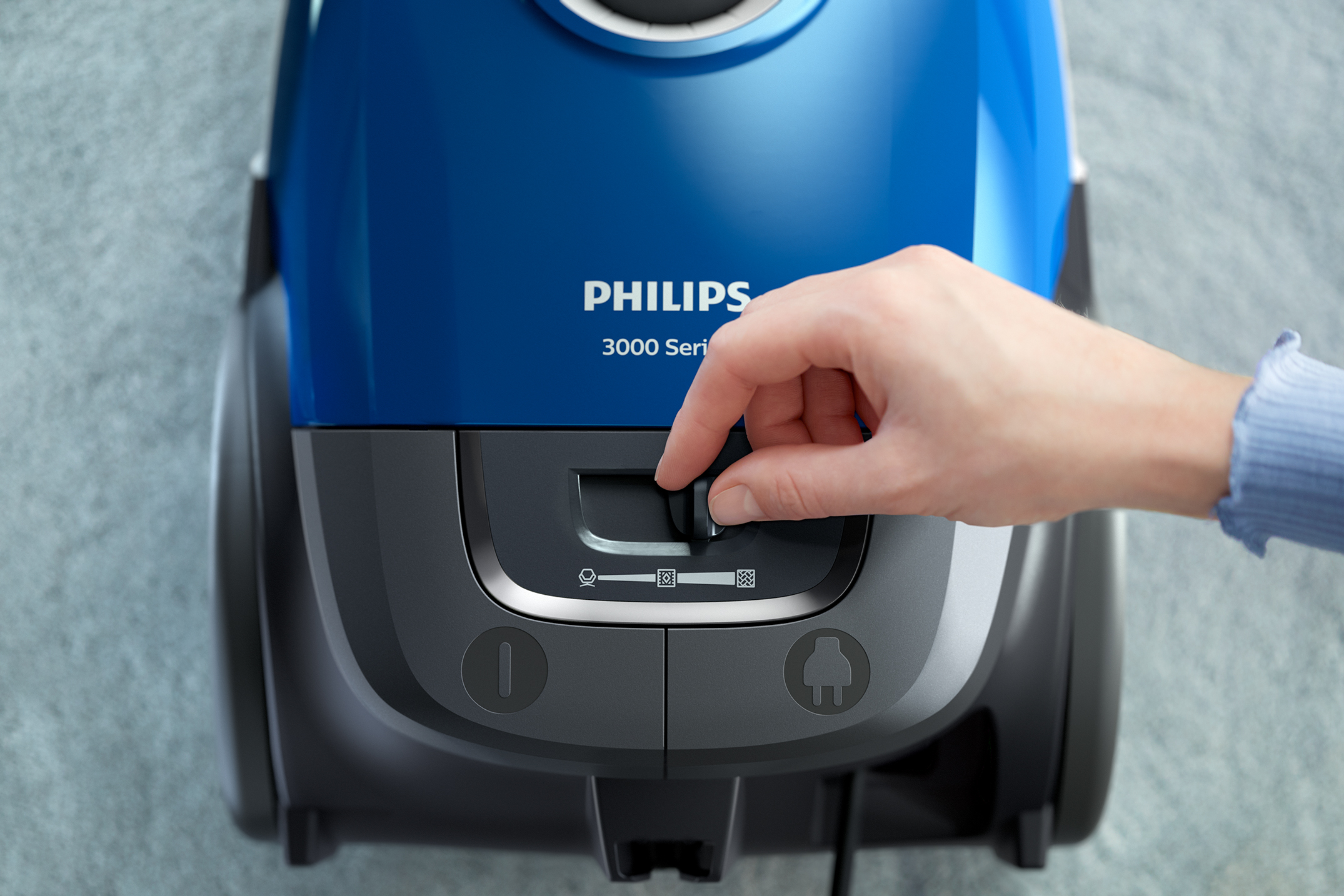 Eine Hand stellt einen Drehregler an einem blauen Philips Staubsauger ein.