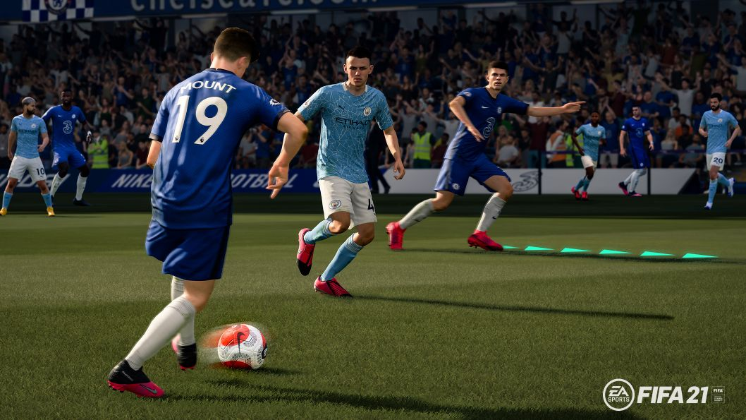 Gameplay de FIFA 21. Jugadores con uniformes azules y celeste en un campo verde. Estadio al fondo.