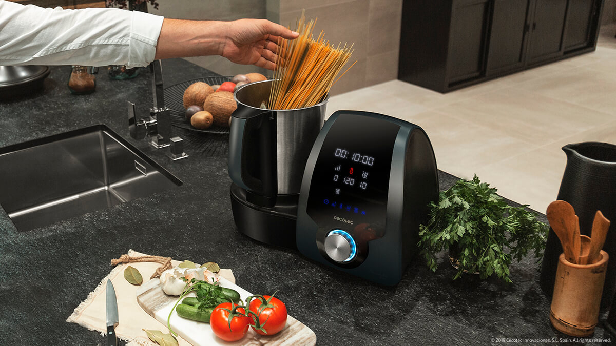 Mano añadiendo espaguetis a una máquina de cocina en una encimera con verduras.
