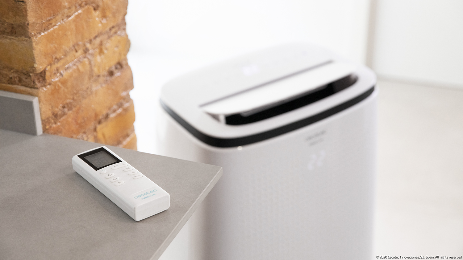 An air conditioner with a remote on a gray surface, near a brick wall. The unit is white.