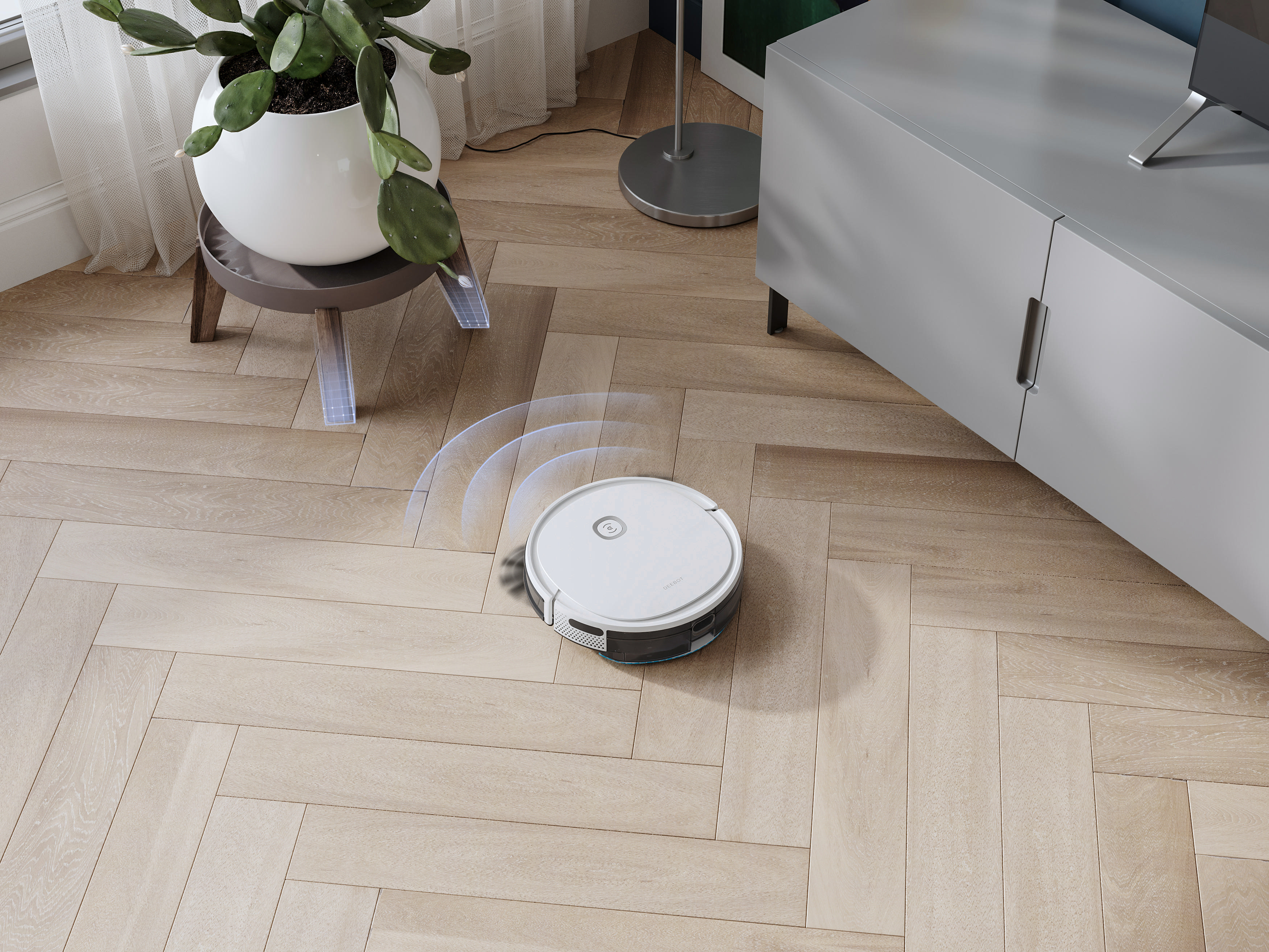 Roboter-Staubsauger auf Holzboden. Links eine Pflanze auf einem Ständer, rechts ein Schrank.