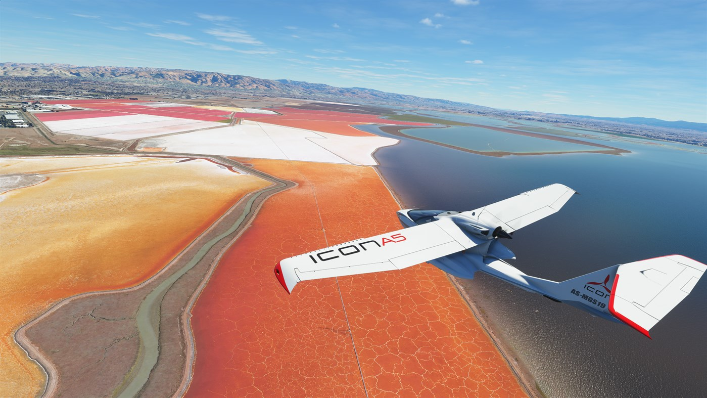 Avion volant au-dessus de marais salants rouges et blancs, avec lac et ville en arrière-plan, ciel bleu.