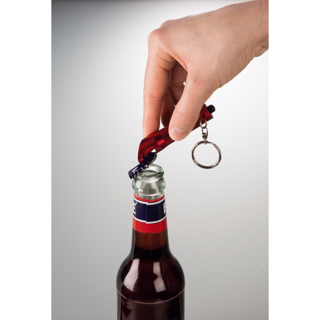 Main utilisant un ouvre-bouteille rouge pour ouvrir une bouteille de liquide rouge, sur fond blanc.