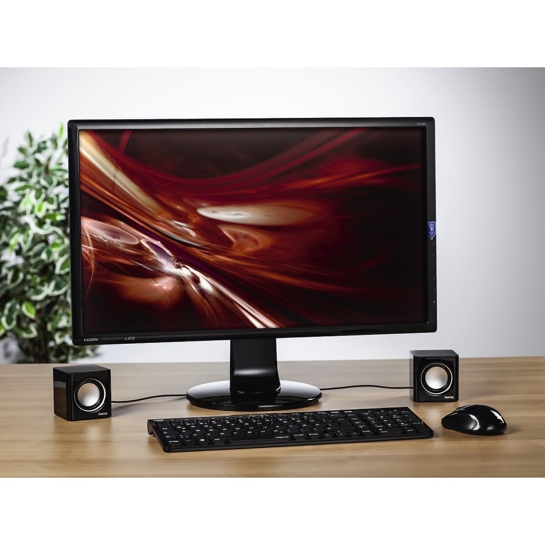 Computeropstelling: monitor, luidsprekers, toetsenbord en muis op een houten bureau. Plant op de achtergrond.