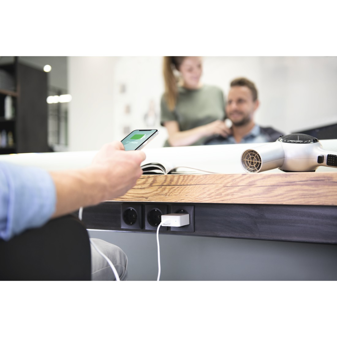 Person using phone at a charging station, with a hairdryer on a counter. People in background.