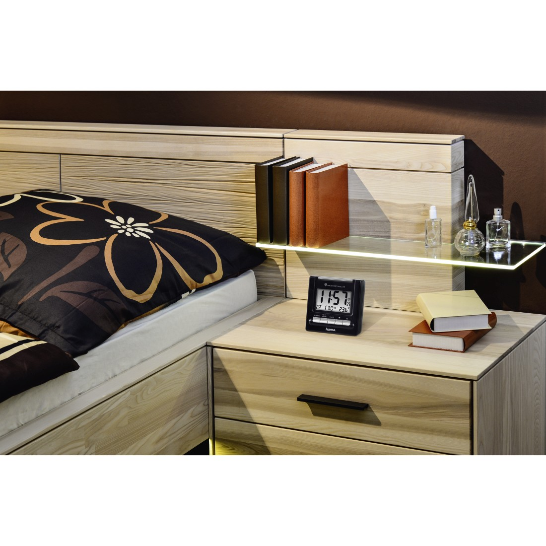 Bedroom with a wooden bed, bedside table with books, clock and perfumes, and a patterned duvet.