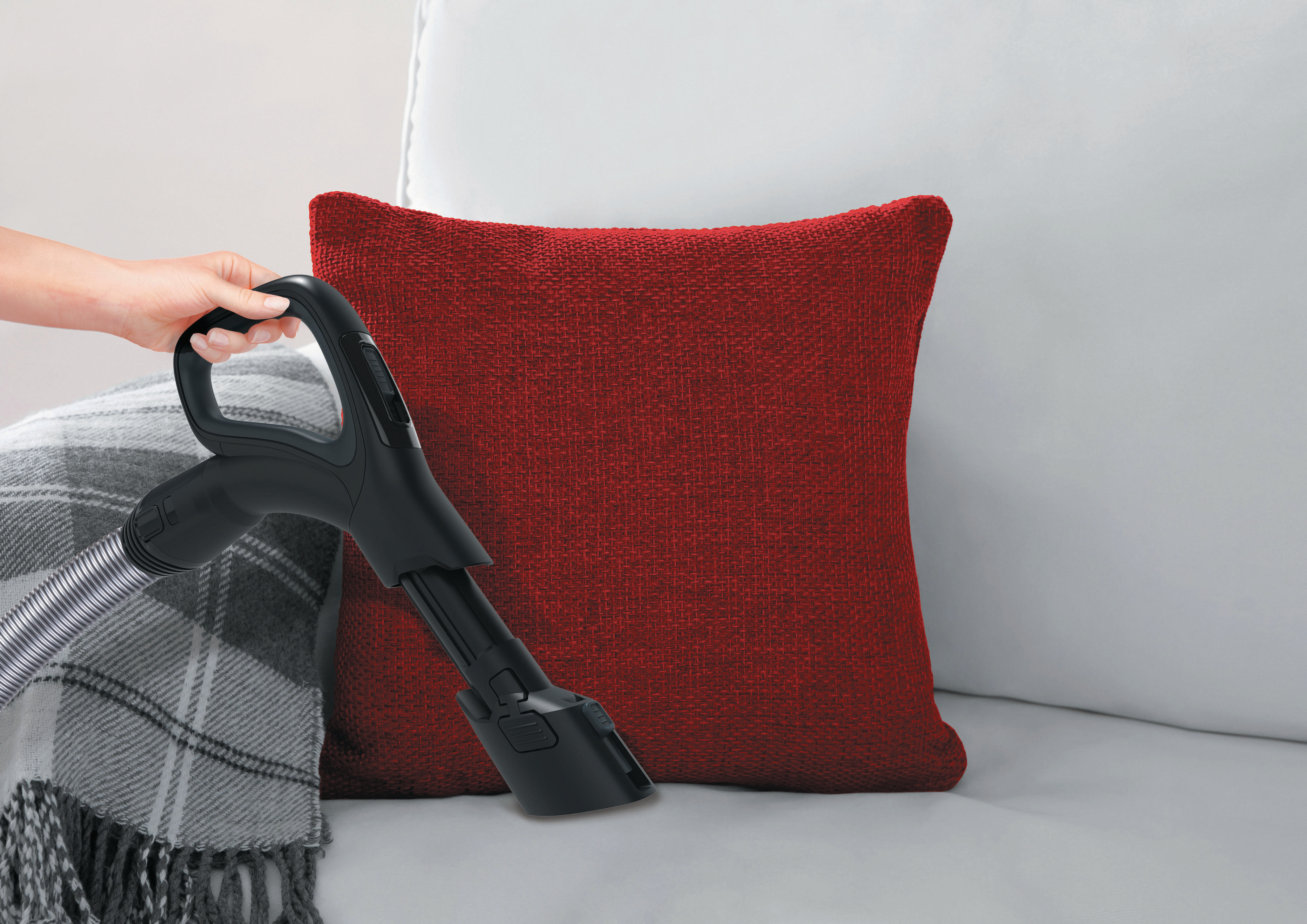 Hand saugt einen roten Kissen auf einem weißen Sofa mit einer grauen Decke ab.