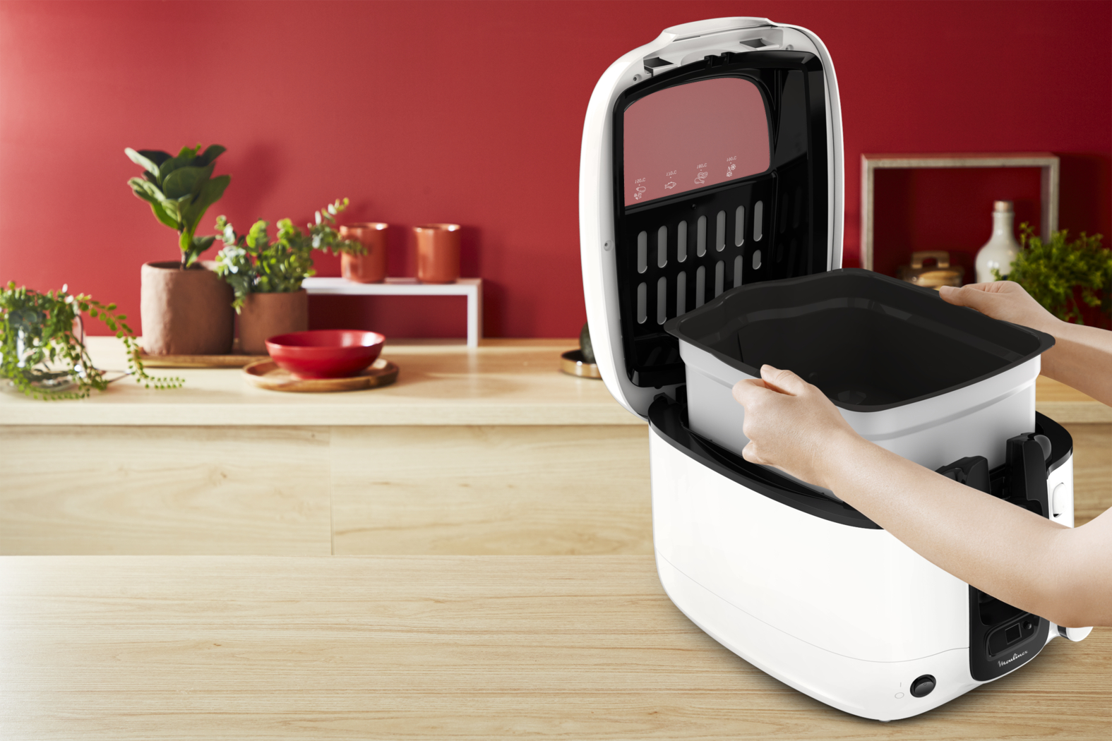 Hands placing a container into a white and black kitchen appliance, with a red wall background.