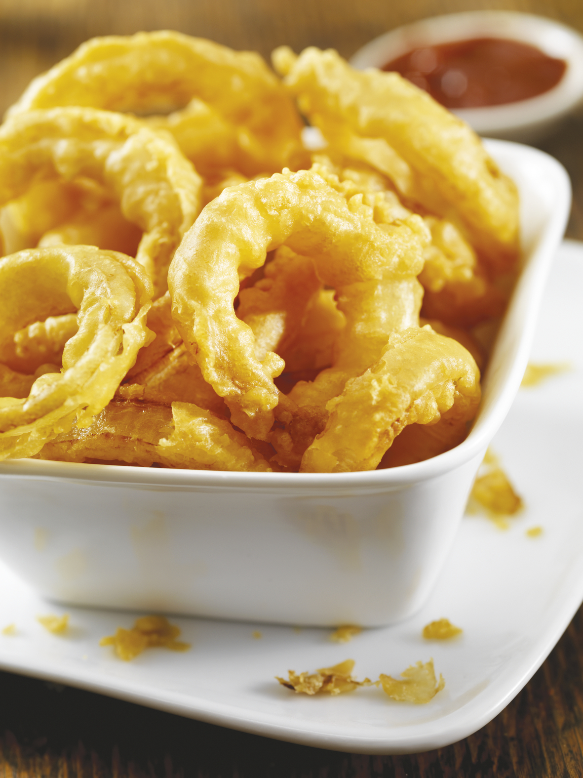 Close up of fried onion rings in a white bowl, with a red sauce in the background.