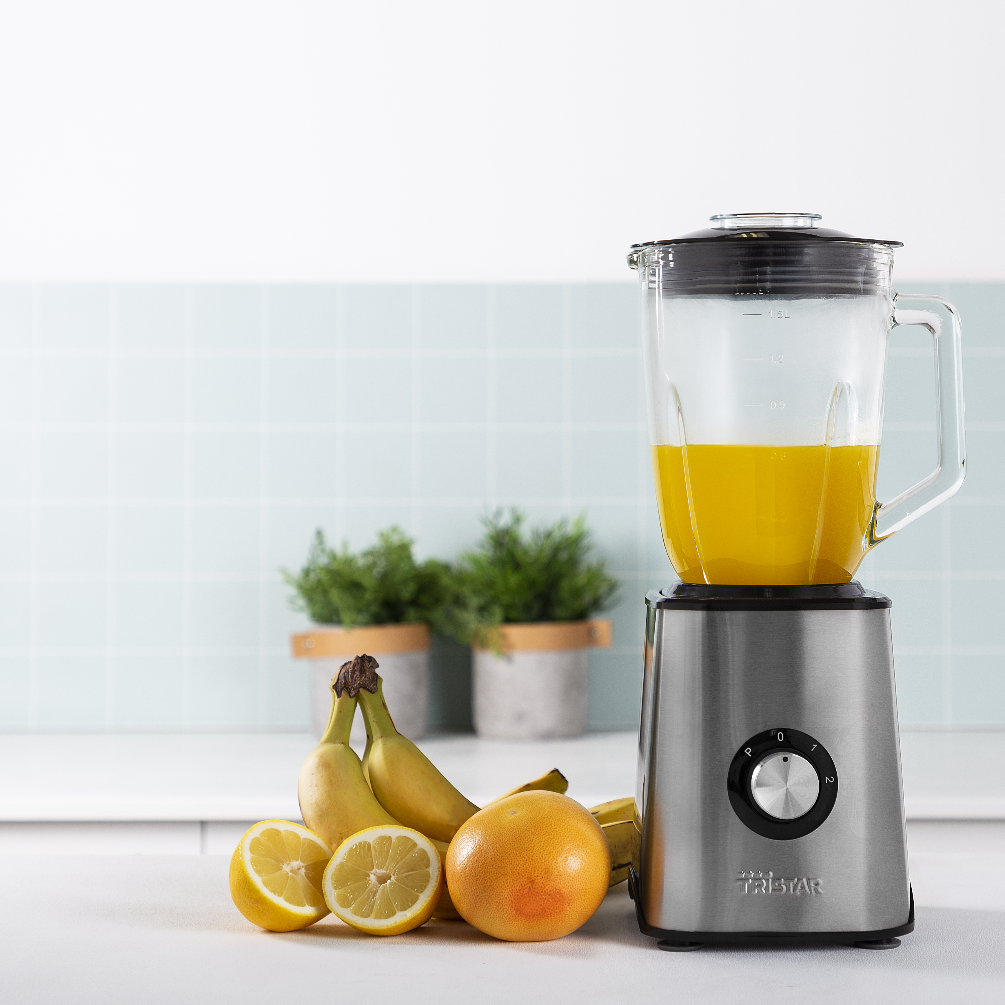 Blender met oranje vloeistof. Bananen, halve citroenen en een grapefruit ervoor. Lichtblauwe achtergrond.
