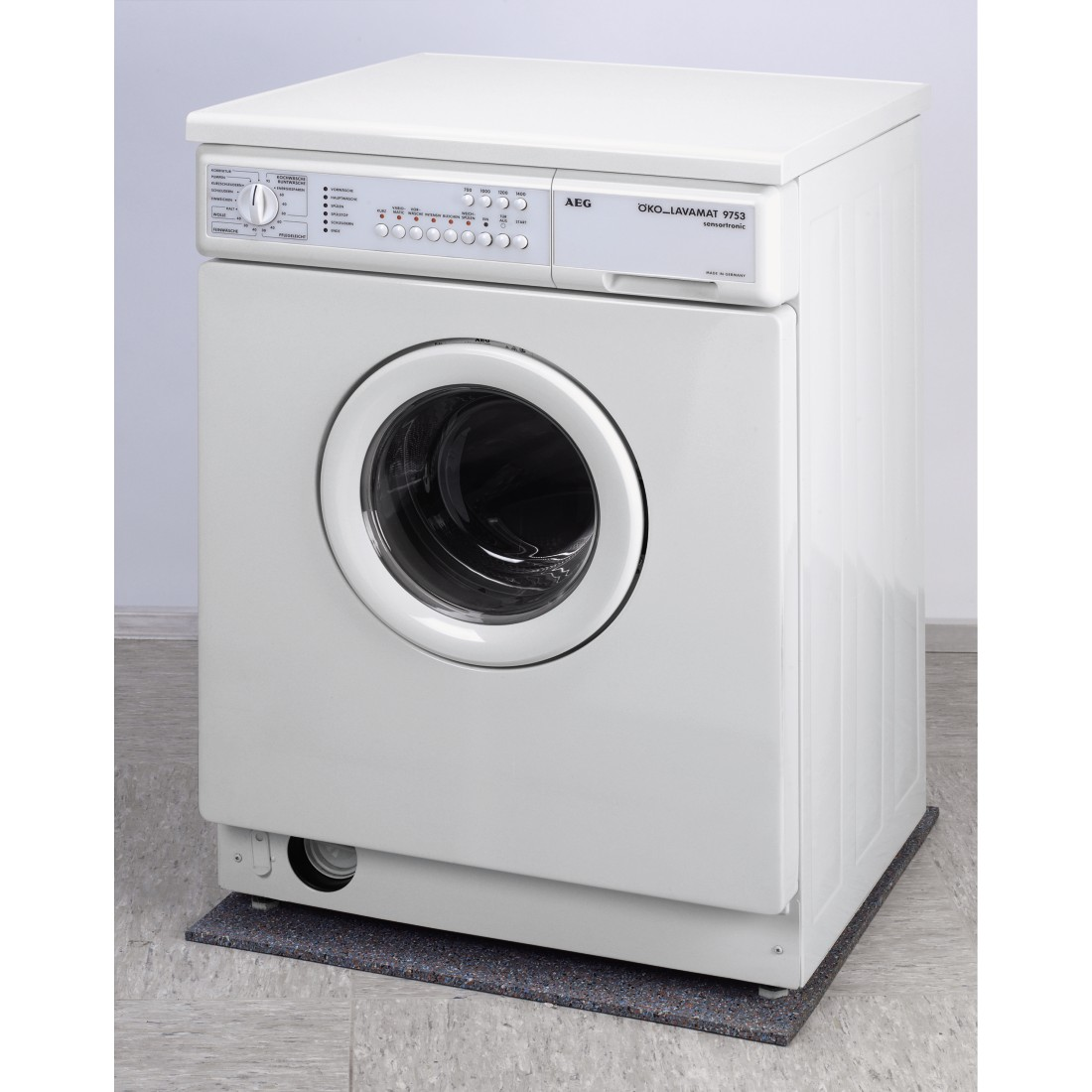 A white AEG washing machine on a gray floor. It has a round door and control panel. It rests on a dark mat.