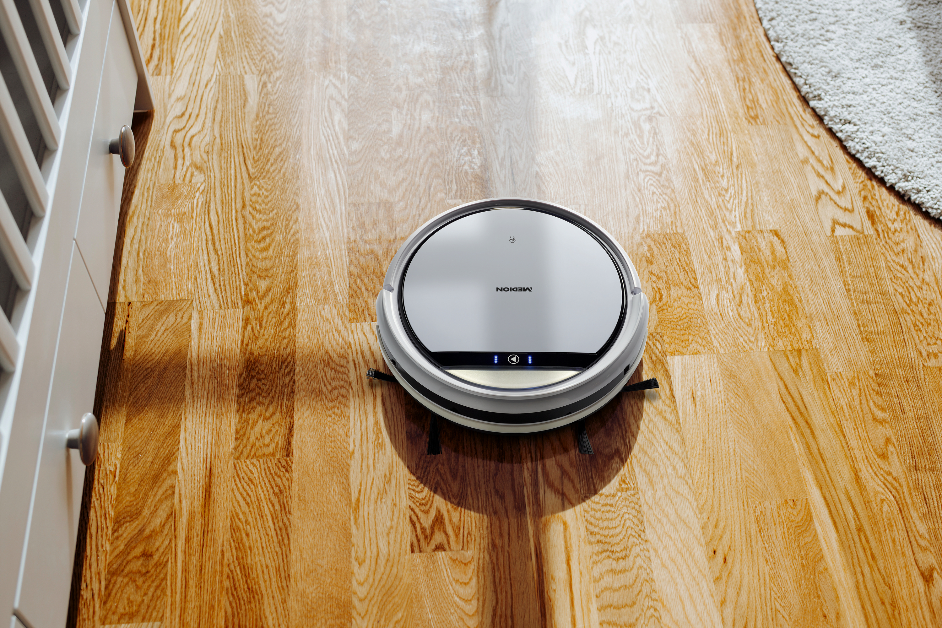 Roboter-Staubsauger auf Holzboden neben weißem Kinderbett.