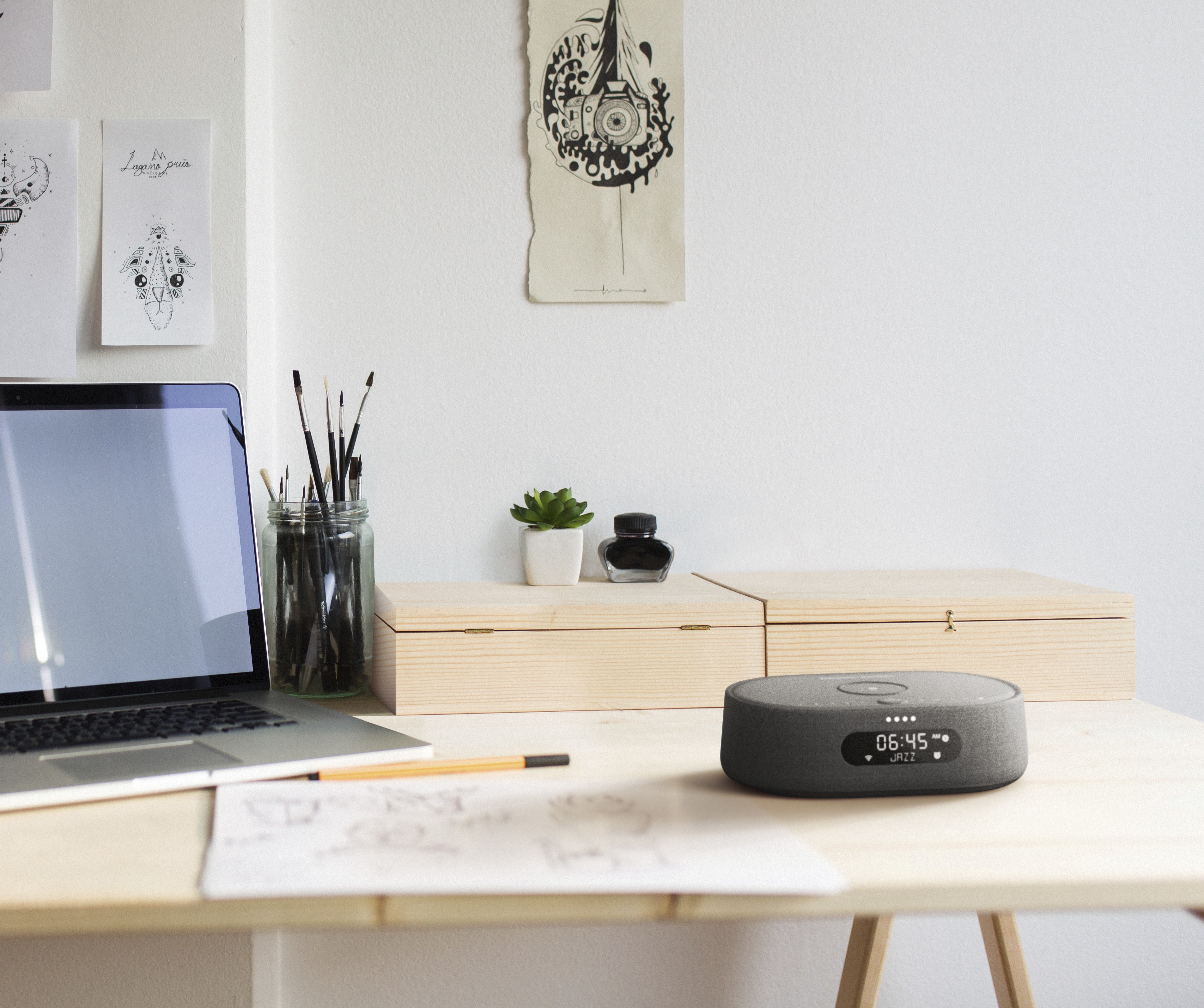 Werkruimte met een laptop, tekeningen en een grijze luidspreker op een houten bureau.