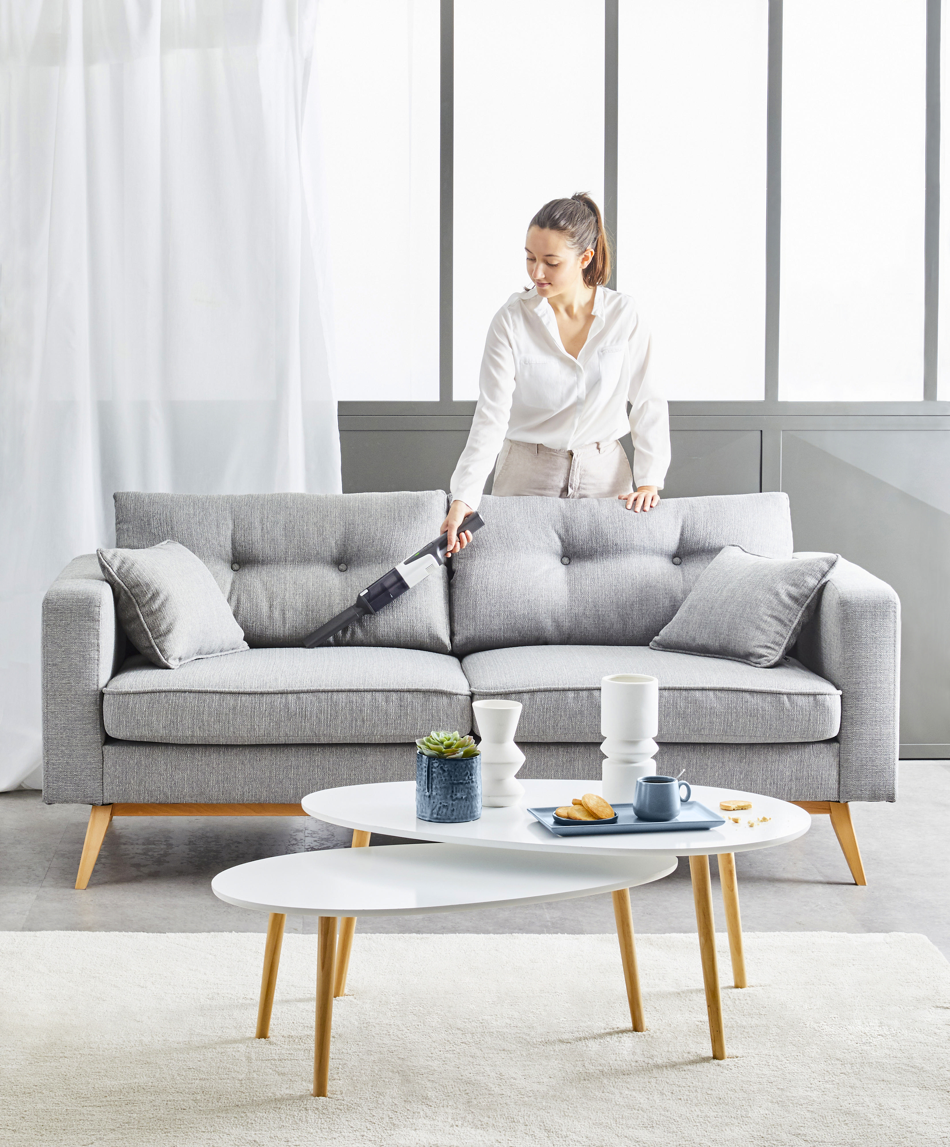 Frau, die ein graues Sofa mit weißen Couchtischen und Dekor staubsaugt.