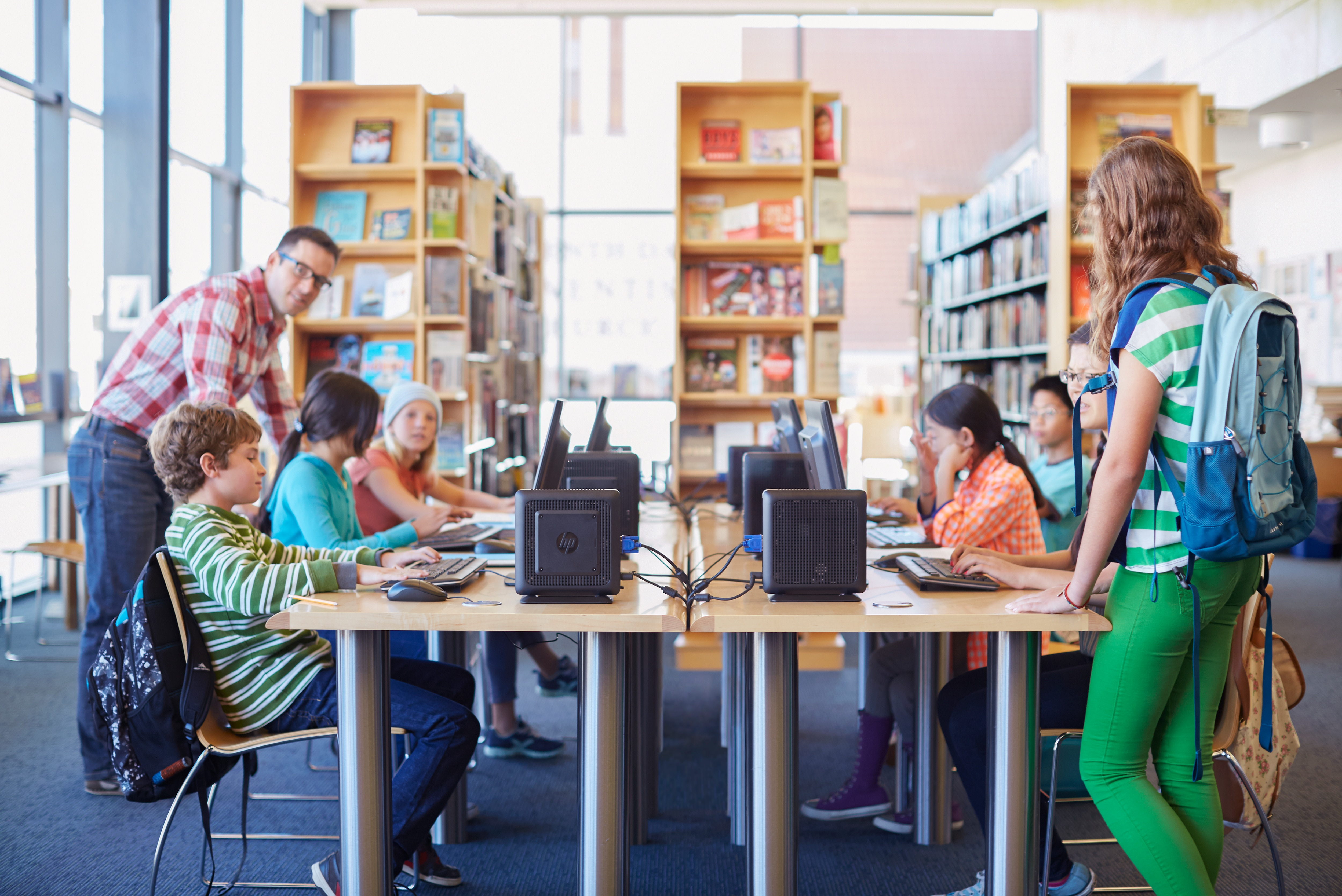 Kinder in einer Bibliothek, die Computer benutzen, mit einem Lehrer. Bücherregale im Hintergrund.