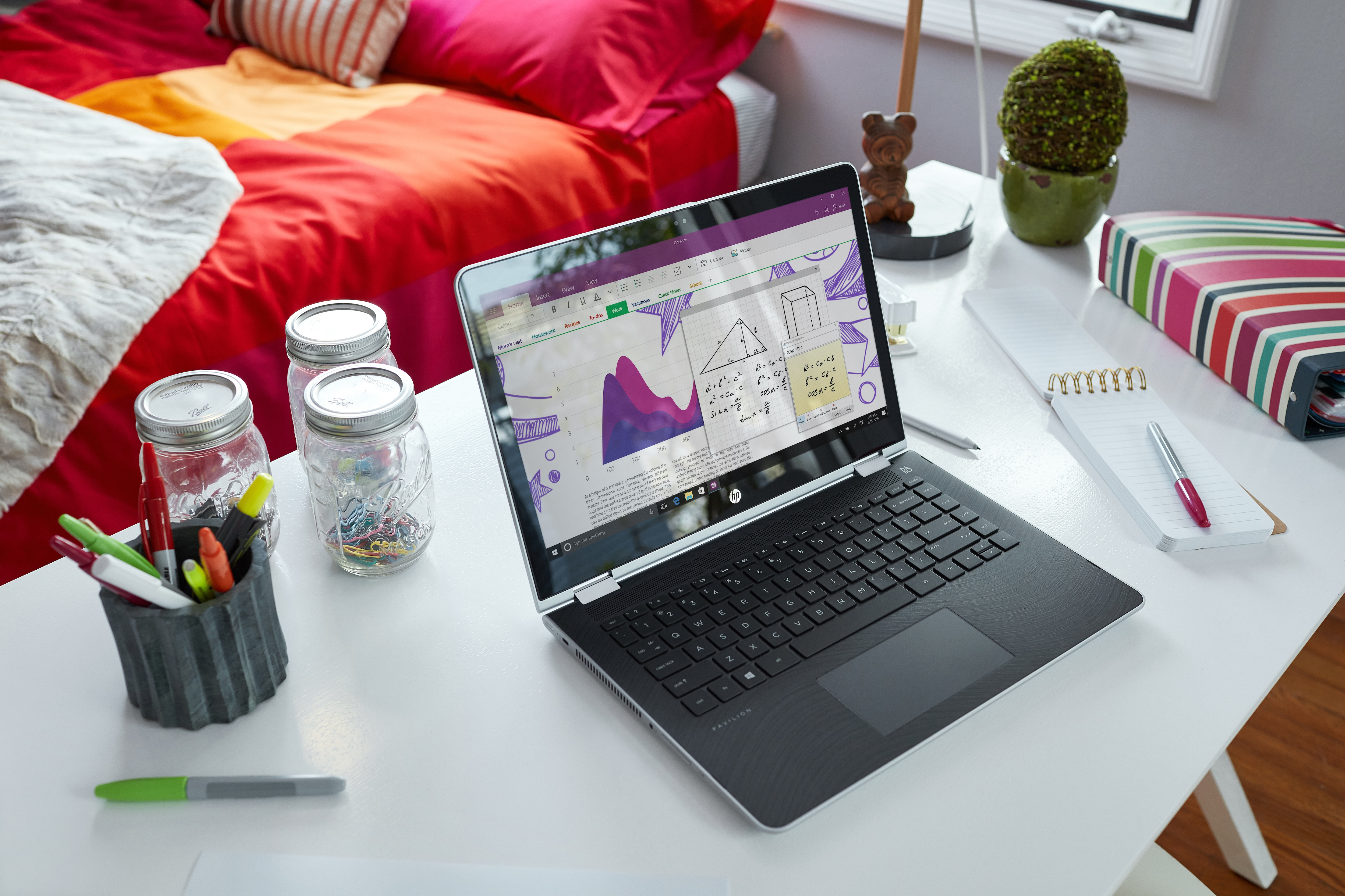 Ein Laptop zeigt Mathe-Notizen auf einem Schreibtisch mit Gläsern, Stiften und einem farbigen Bett im Hintergrund.