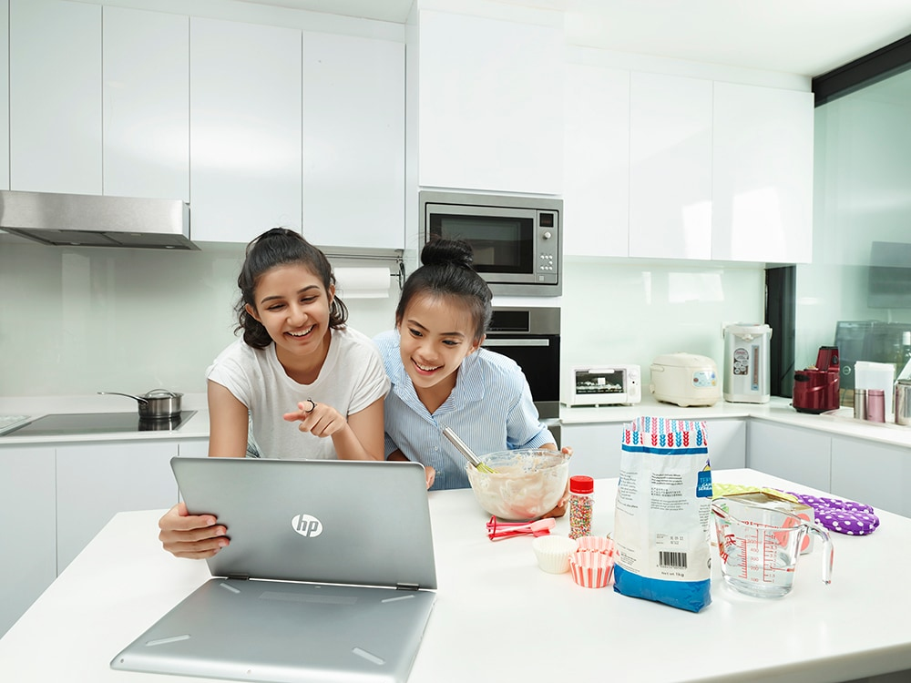 Zwei junge Frauen in einer modernen weißen Küche lachen, während sie auf ein Laptop schauen und backen.
