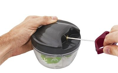 Manos usan un picador de alimentos manual, dentro hay verdura picada. Se adjunta un cordón rojo.