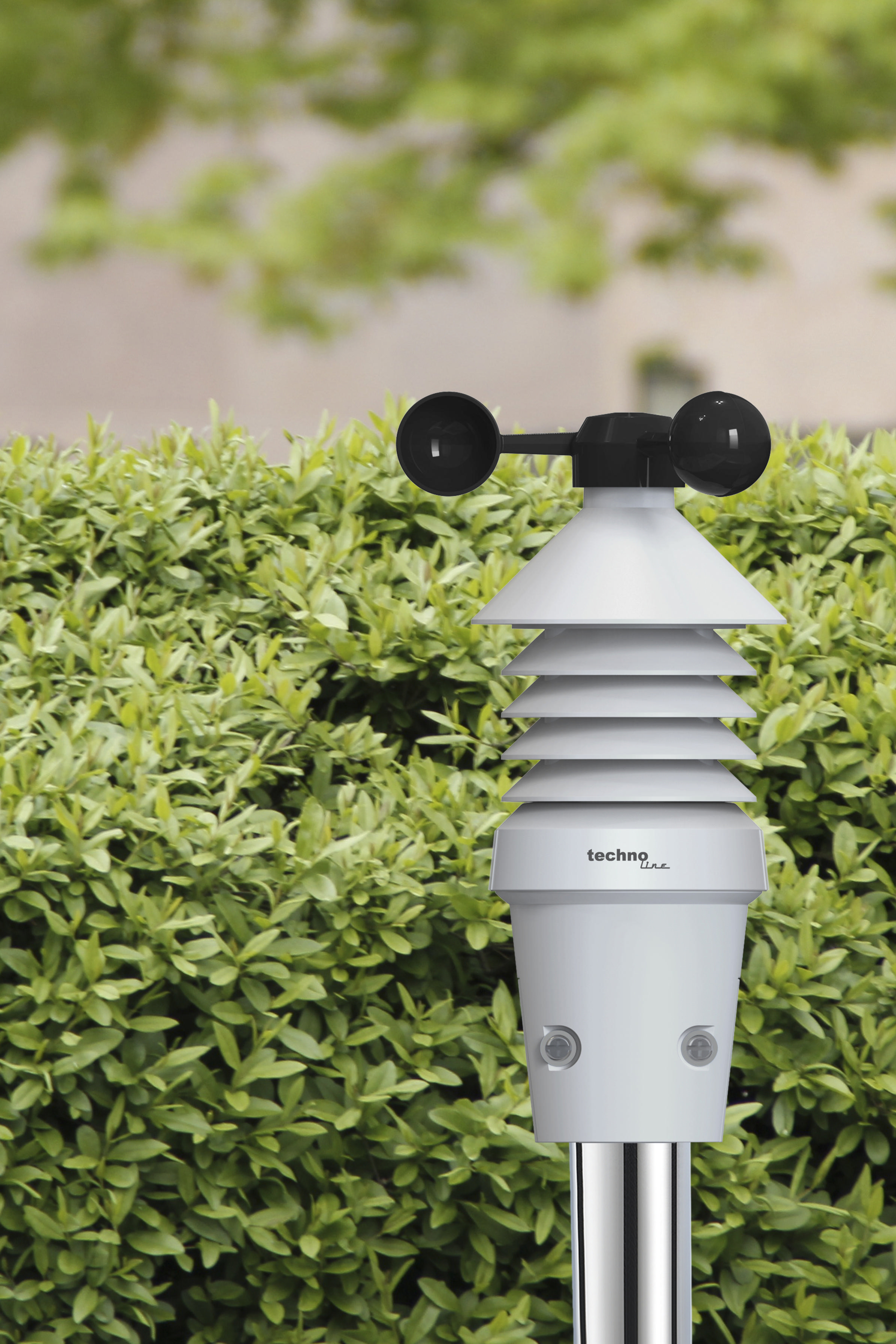 Eine weiße Wetterstation mit schwarzen Sensoren steht vor einer grünen Hecke.