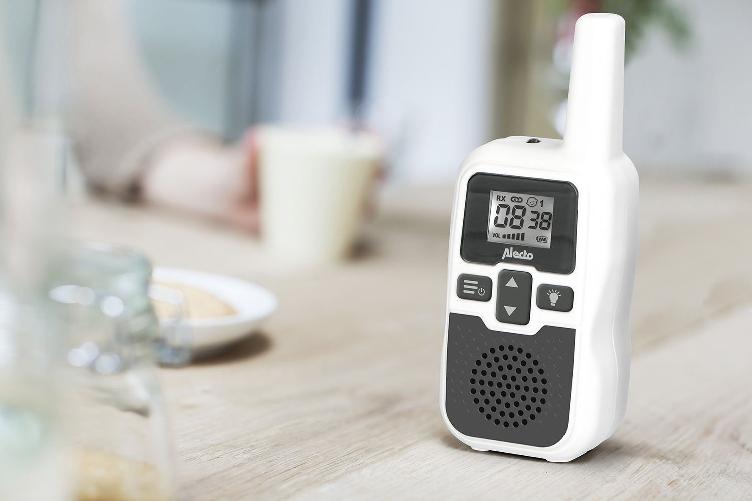 Een witte walkie-talkie op een houten tafel, met een gedeeltelijk zichtbare beker en pot op de achtergrond.