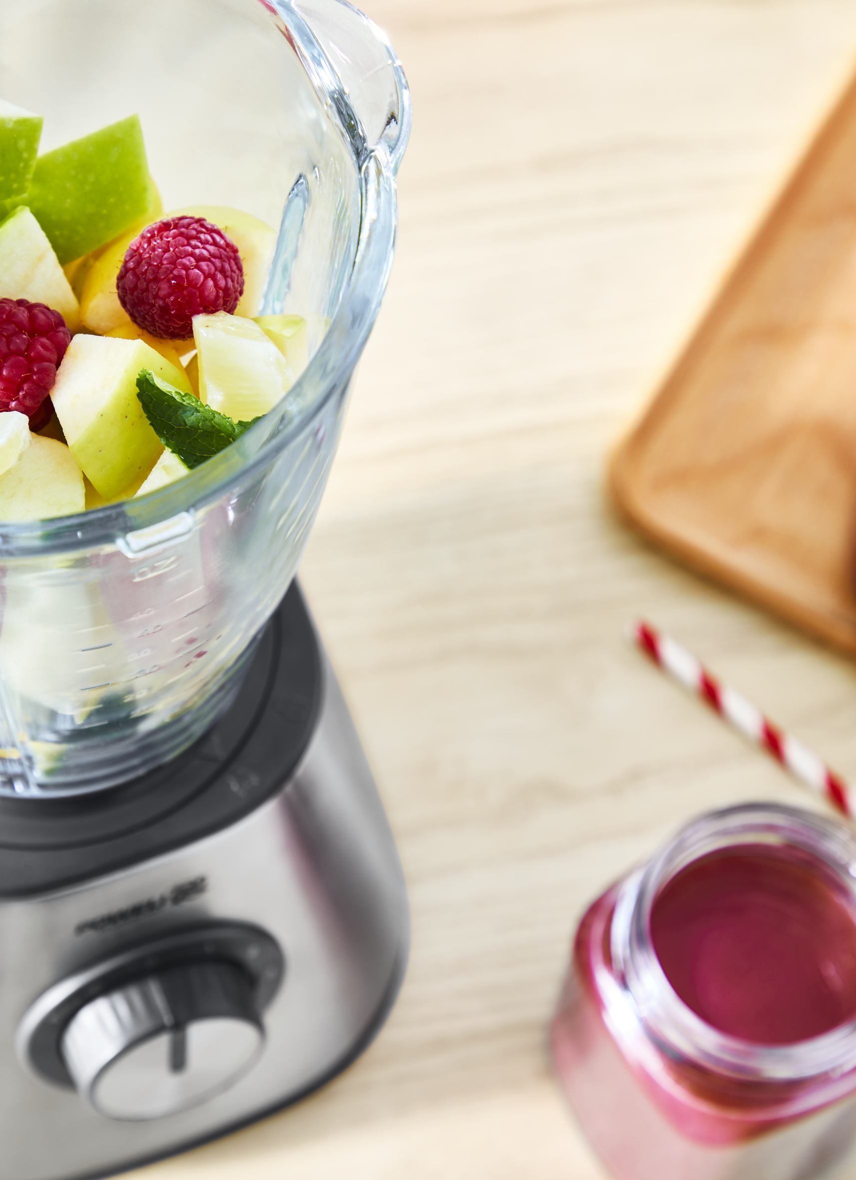 Blender with fruits, raspberries and apple slices. Jar of red liquid and straw.