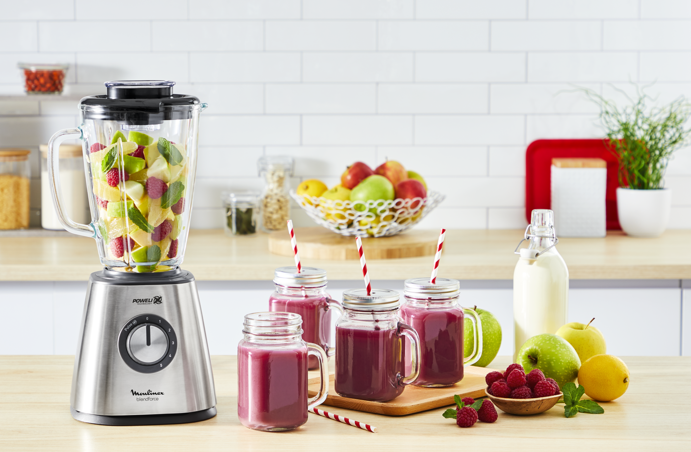 A blender with fruits and smoothies in jars on a wooden board. Straws and fruit are present.